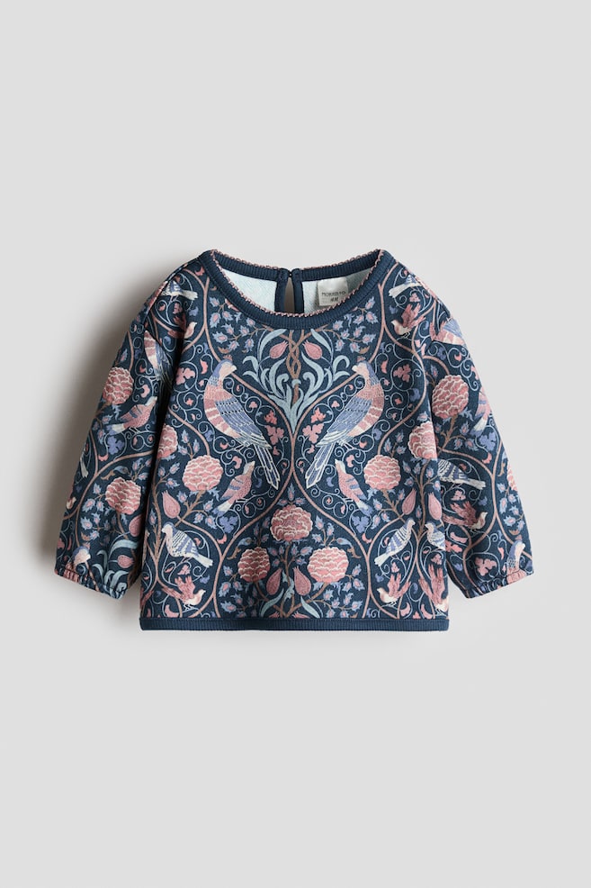 Un sweat en coton lÃ©ger est prÃ©sentÃ© de face, avec un imprimÃ© symÃ©trique sur lâensemble composÃ© dâoiseaux et de motifs floraux dans des nuances de rose et de bleu sur un fond bleu marine foncÃ©. Le vÃªtement prÃ©sente des Ã©paules tombantes, des manches longues avec des poignets Ã©troits Ã©lastiquÃ©s, un col rond bordÃ© de picots et une finition cÃ´telÃ©e Ã  lâourlet. - 1