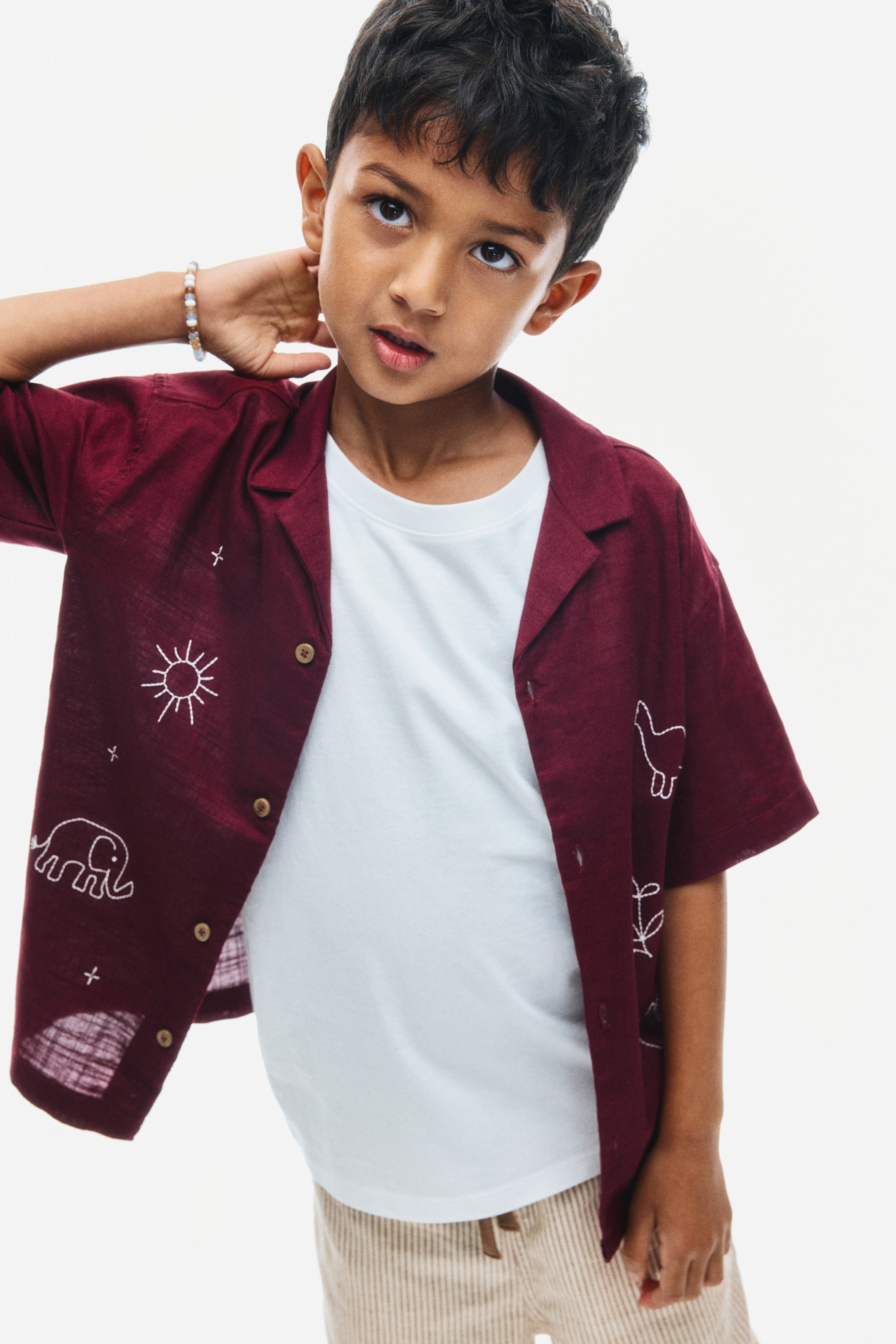 View larger image: A burgundy, short-sleeve shirt in a textured cotton weave is worn open over a white T-shirt, showcasing its relaxed fit and dropped shoulders. The front-facing view highlights its resort collar, brown buttons, and white embroidered line-art designs, including an elephant, a sun, and stars.
