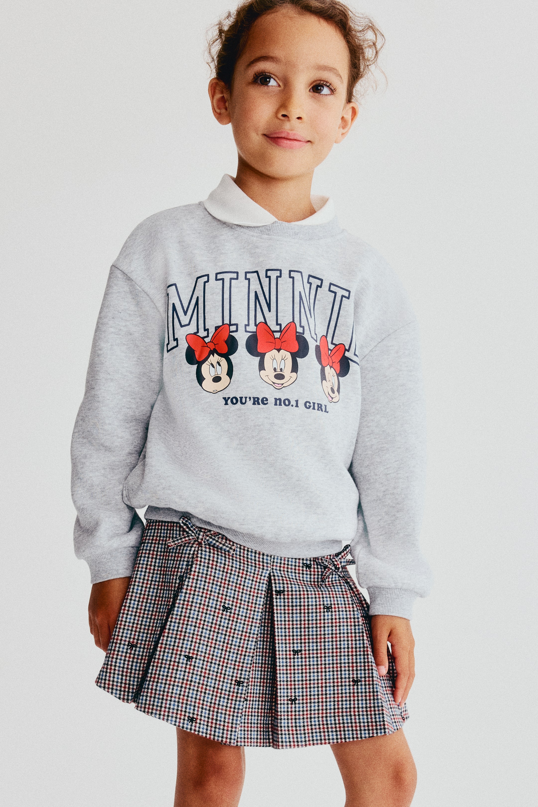 View larger image: A light gray crewneck sweatshirt is shown from the front, featuring dropped shoulders, ribbed trims, and a printed graphic of the word "MINNIE" above three Minnie Mouse characters and the text "YOU'RE NO. 1 GIRL." The sweatshirt is worn layered over a white collared shirt.