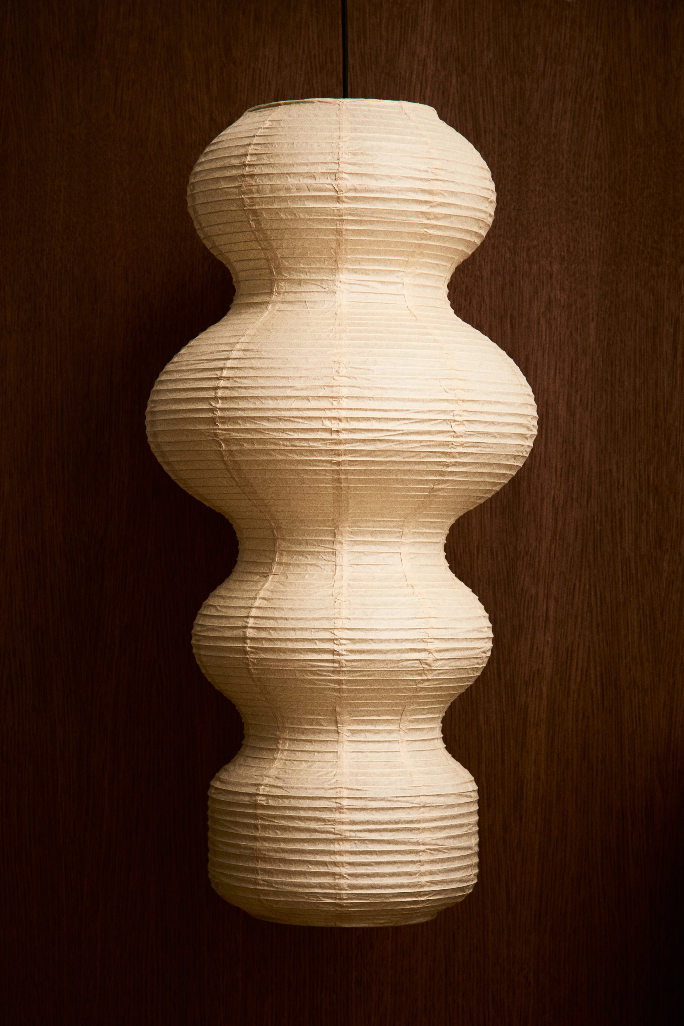 Shown from the front, a pendant light featuring a multi-tiered, sculptural rice paper lampshade in a natural beige hue is hanging from a thin black cord. The lampshade displays a textured, horizontally ribbed surface with four distinct bulbous segments of varying widths.
