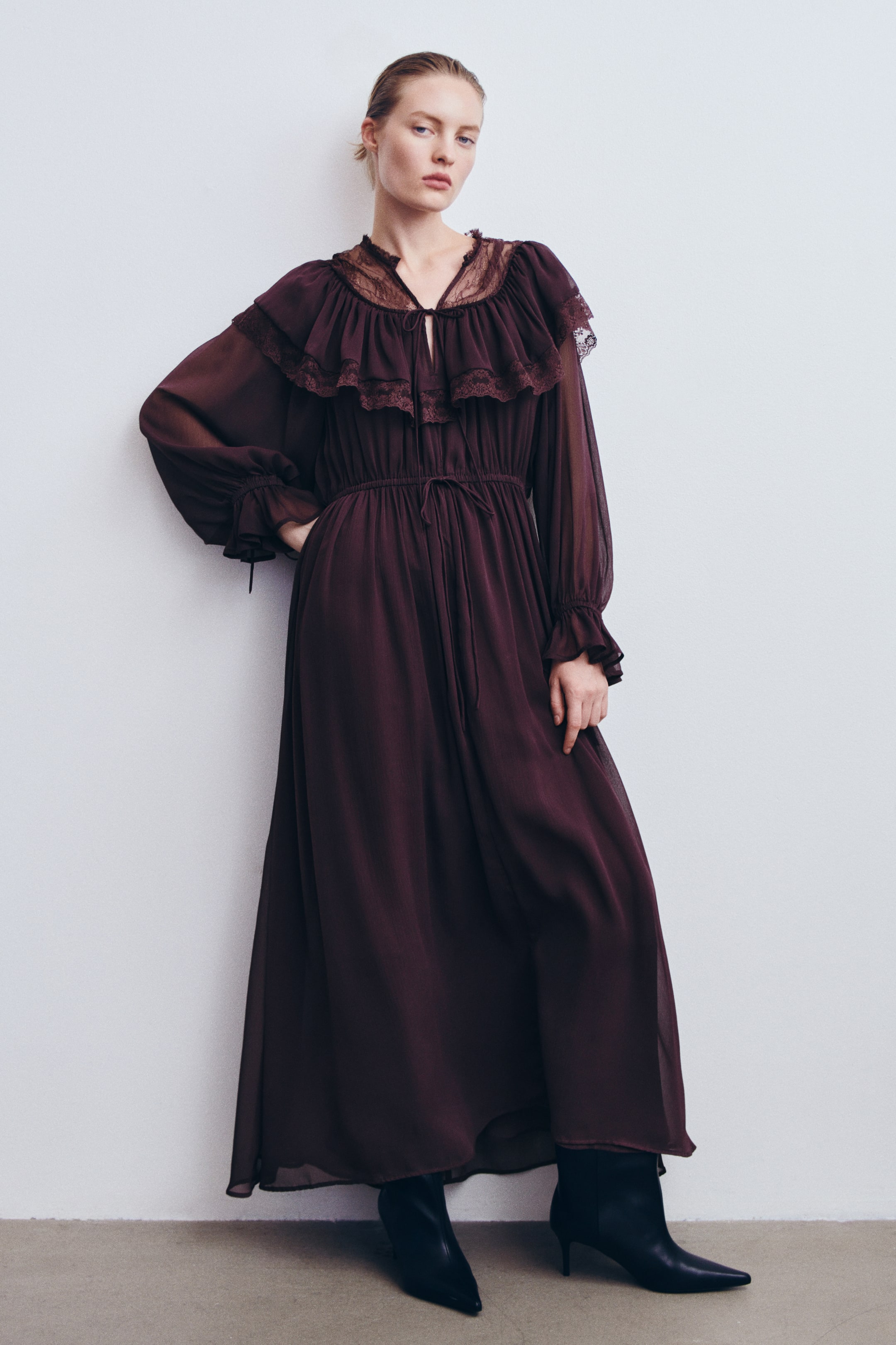 Visa större bild: En mörk plommonfärgad maxiklänning i krinklad chiffon bärs av modellen, med ett volangprytt ok med spetskant runt V-ringningen och genomskinliga ballongärmar med elastiska manschetter. Klänningen har dragsko i halsen och midjan och stylas med svarta högklackade stövletter med spetsig tå som syns vid fållen.