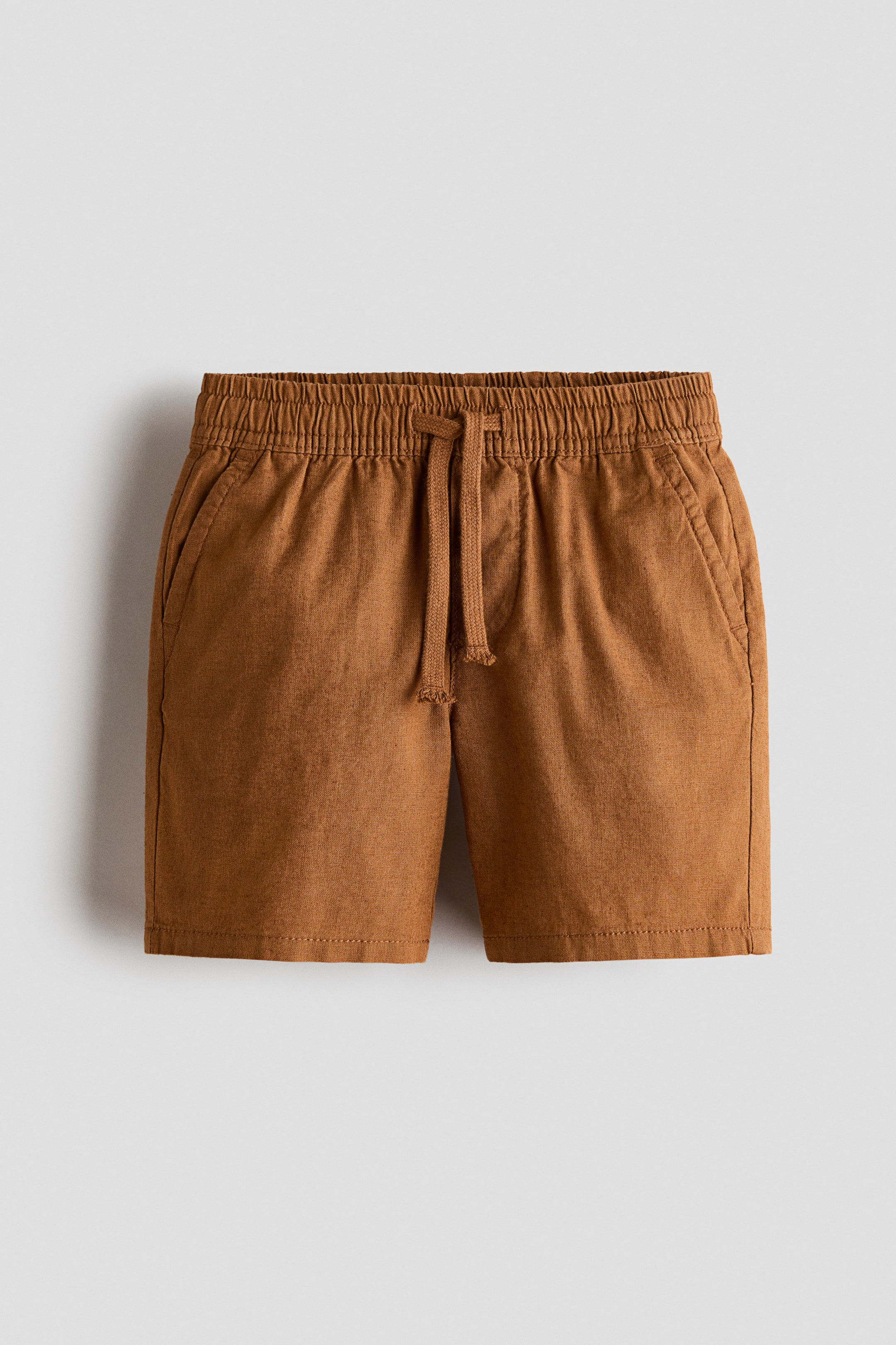 Xem ảnh lớn hơn: Một chiếc quần short màu nâu, chất liệu pha linen và cotton, được chụp chính diện, với cạp chun kèm dây rút cùng màu và chi tiết khóa giả. Thiết kế có túi chéo phía trước và được làm từ chất vải dệt có kết cấu, thoáng nhẹ.