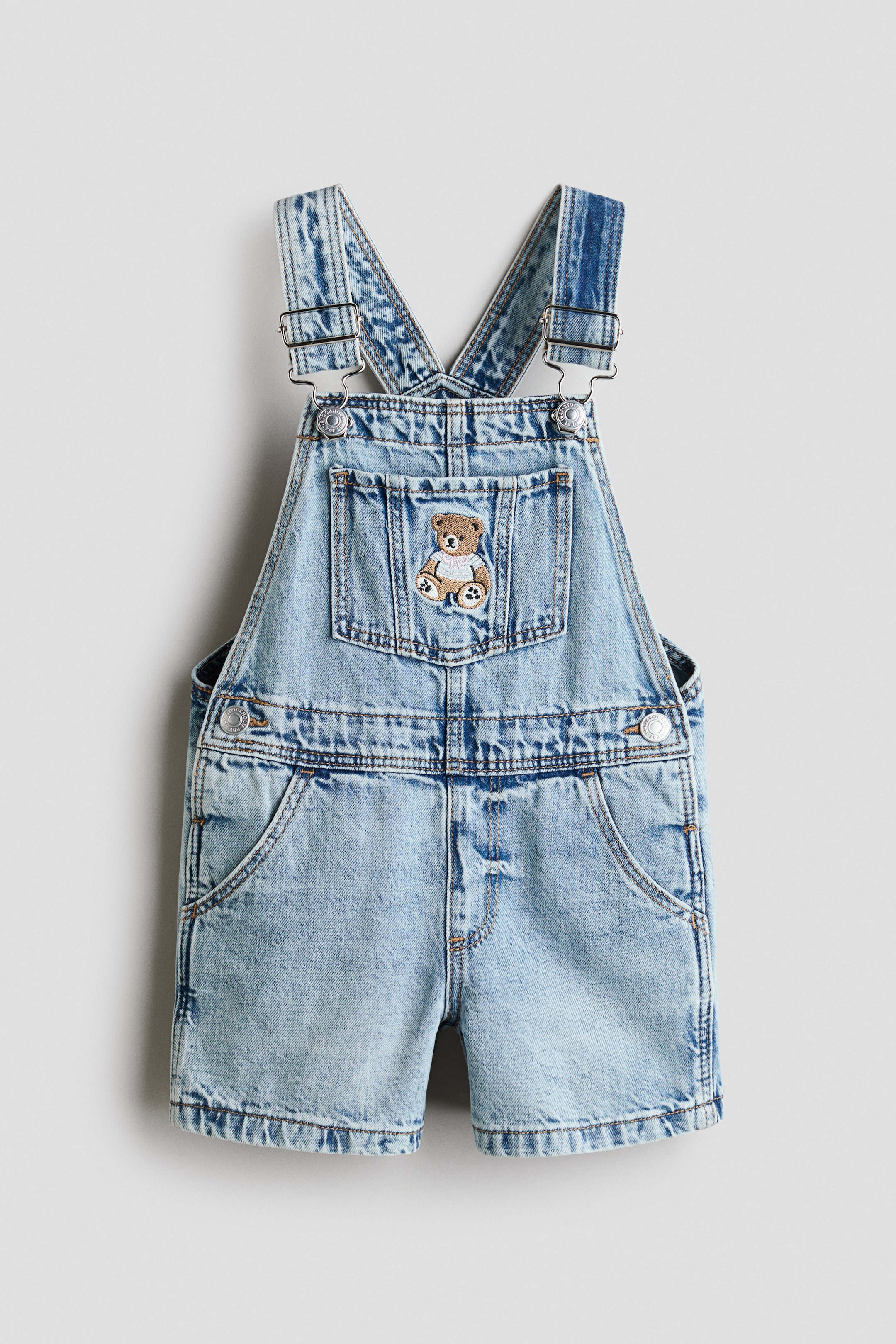 View larger image: Denim short overalls are facing front, featuring a light blue acid-wash finish with adjustable shoulder straps and silver-colored buckles. A small embroidered brown teddy bear wearing a white shirt is decorating the front bib pocket, and front slash pockets are visible.