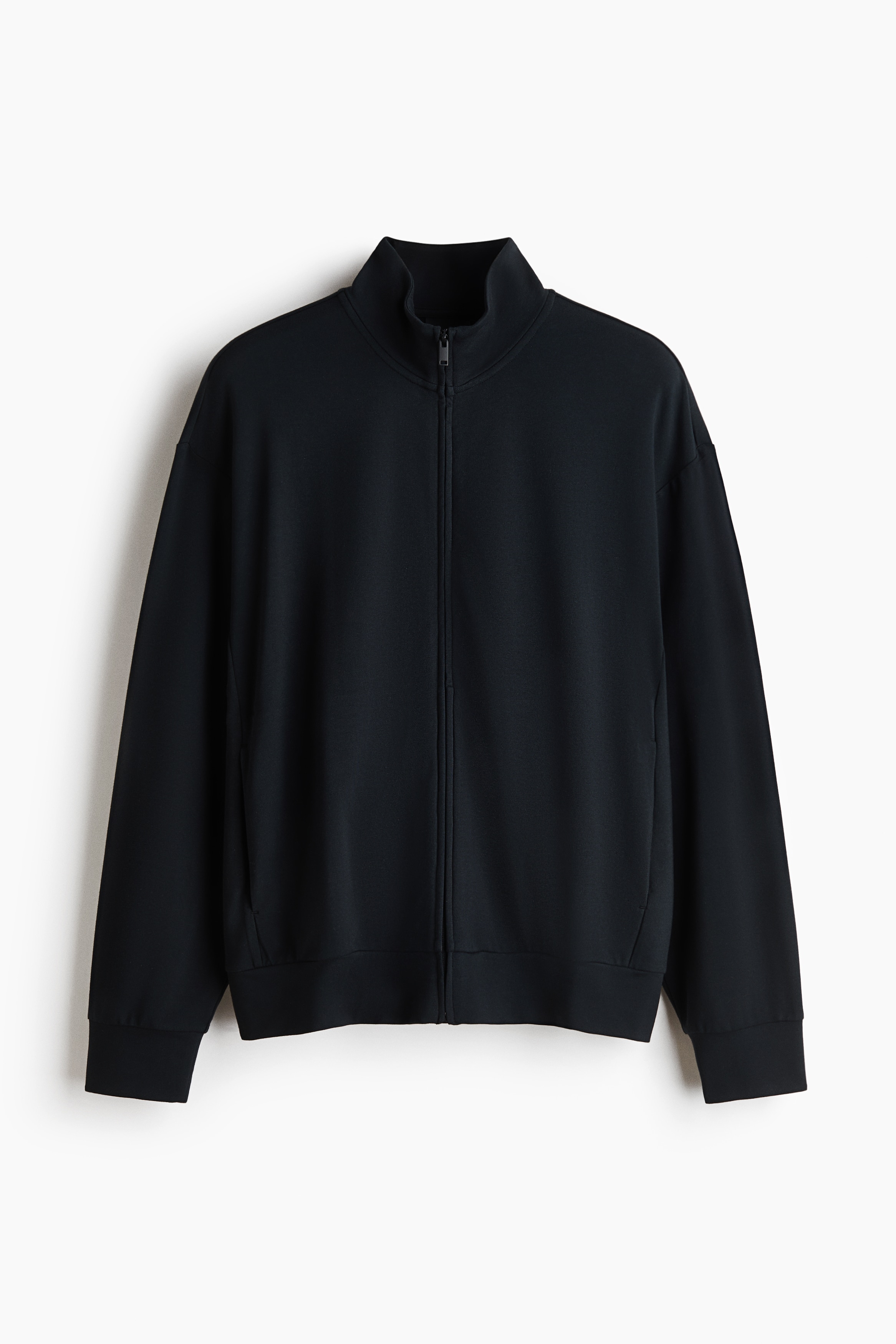 Une veste en tissu molletonnÃ© noir avec col montant, fermeture Ã©clair Ã  lâavant et Ã©paules tombantes est portÃ©e par le mannequin. La veste coupe dÃ©contractÃ©e est agencÃ©e avec un pantalon Ã  cordon de serrage gris foncÃ©.