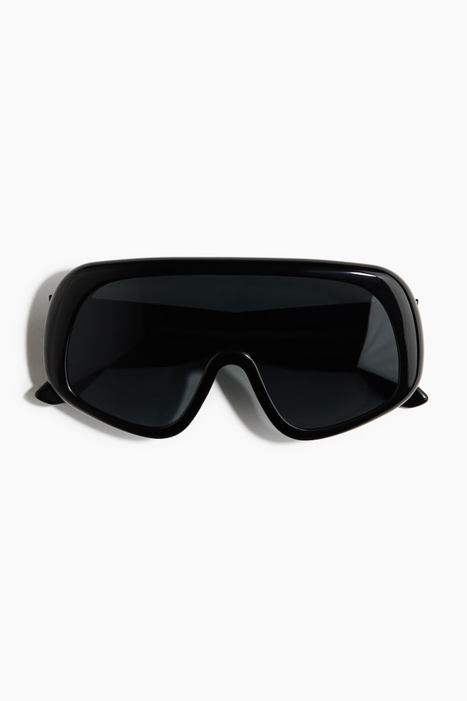 The black shield-style sunglasses feature a wide, dark lens seamlessly integrated into a thick, full-rim frame. The frame curves around the sides, creating a wrap-around design. - 1