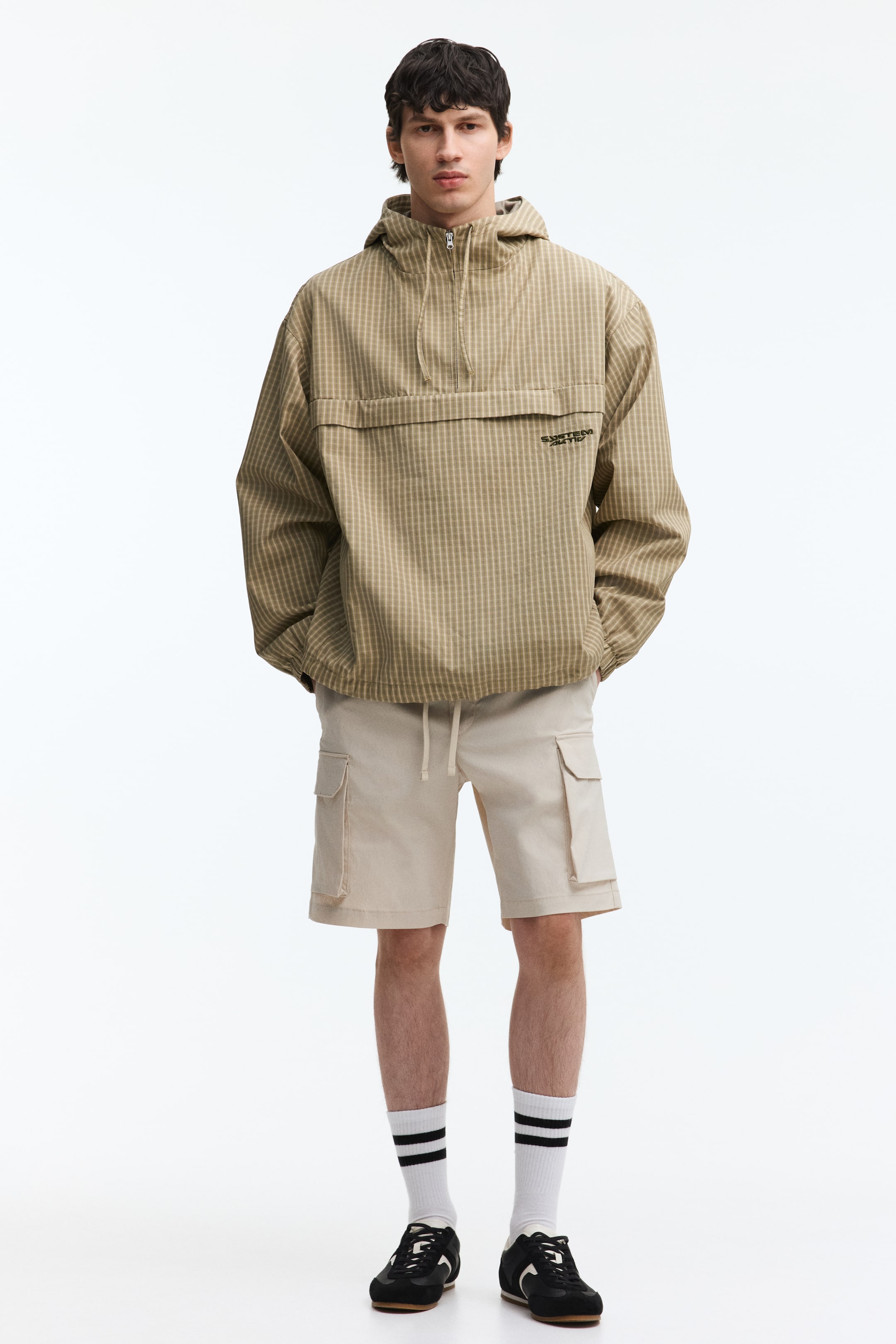 View larger image: A beige anorak jacket with a subtle grid pattern is facing forward. It features a hood, a half-zip front, and a large horizontal flap pocket across the chest, displaying "SURFER RATIO" embroidered in black.