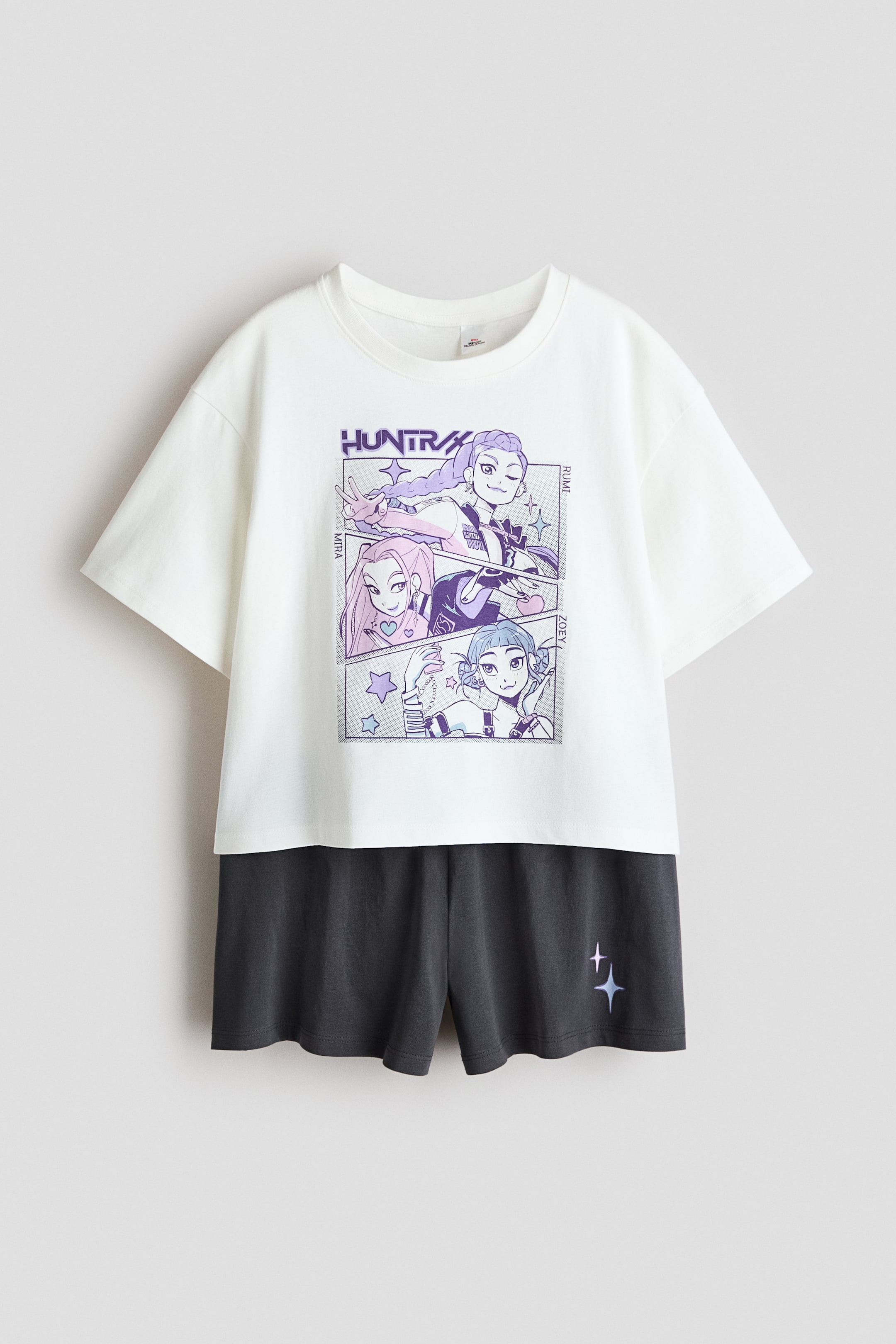 Visa större bild: Ett pyjamasset visar en vit T-shirt och mörkgrå shorts, presenterade framifrån. Den vita T-shirten har en ribbad rund hals, nedhasade axlar och ett grafiskt tryck på framsidan som avbildar tre kvinnliga karaktärer i anime-stil i serierutor, medan de matchande mörkgrå shortsen har en elastisk midja och en liten stjärngrafik på det nedre högra benet.