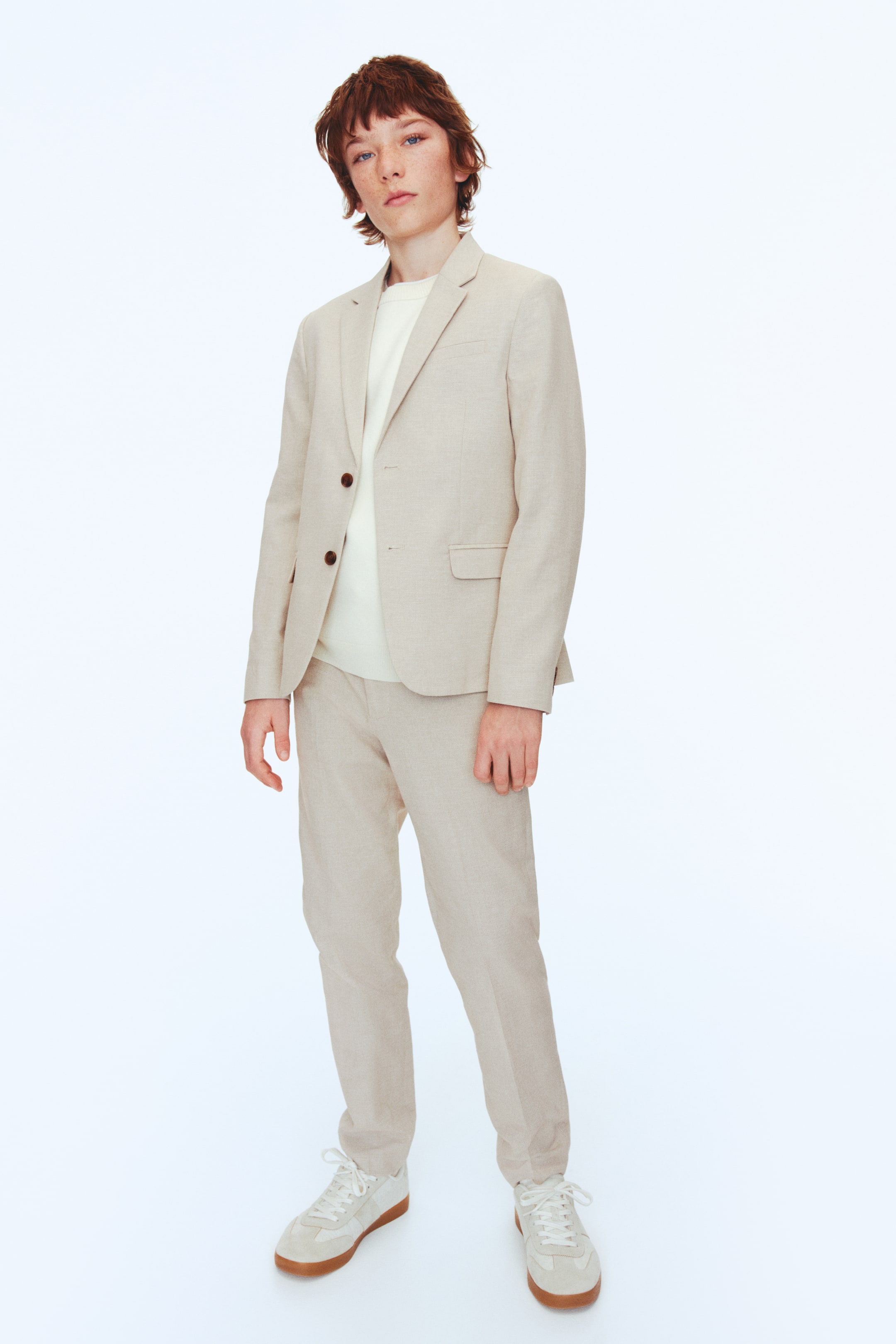 Agrandir l'image: Un costume beige clair, confectionné dans un tissu mélangé de coton et de lin tissé, est présenté de face. La veste classique présente une fermeture à deux boutons avec des boutons foncés, des revers crantés, une poche poitrine factice, des poches avant à rabat, et des boutons décoratifs aux poignets, tandis que le pantalon assorti arbore des poches latérales en diagonale et des jambes fuselées avec des plis.
