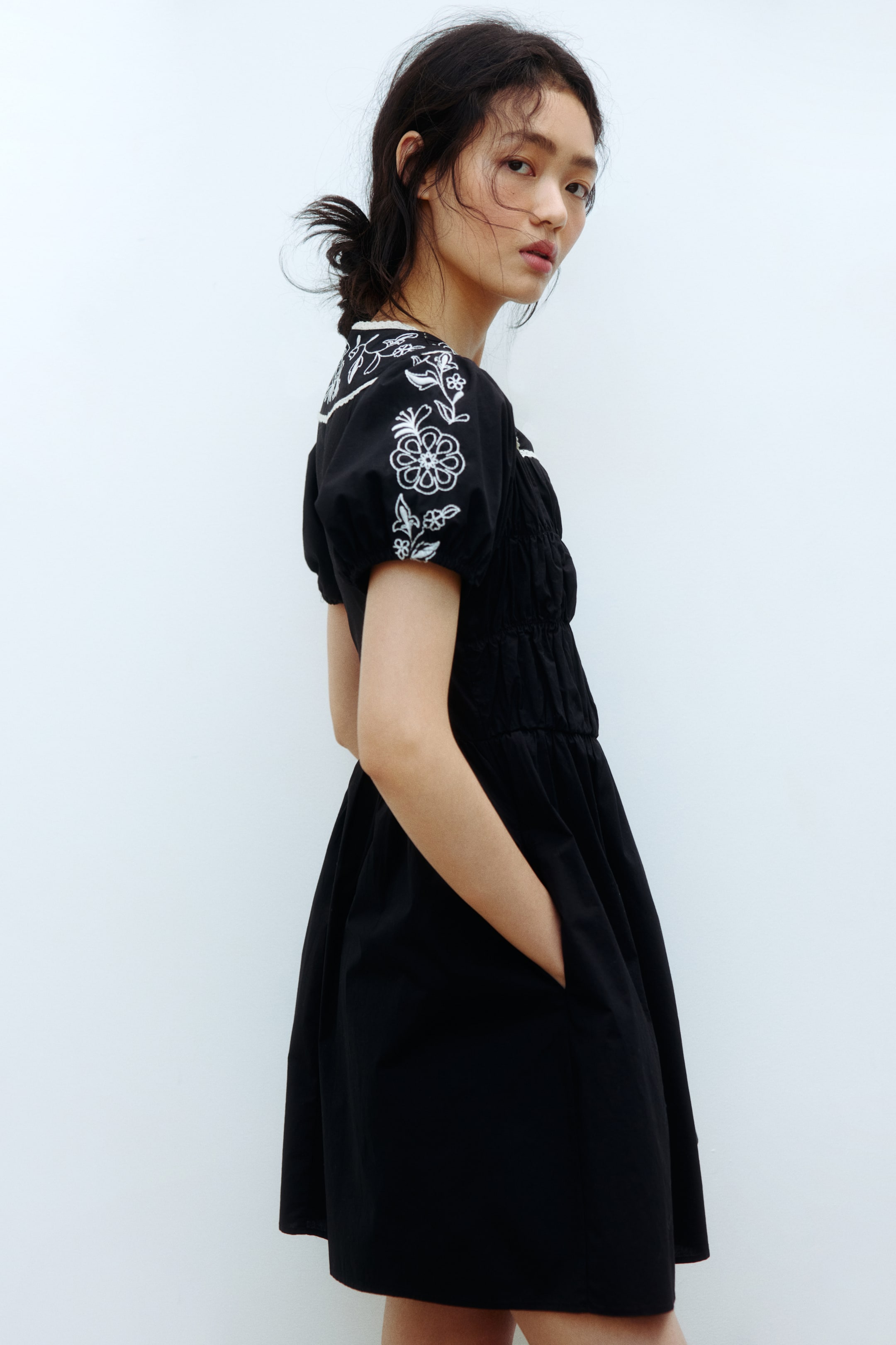 View larger image: A black dress showcases white floral embroidery on its puffed short sleeves and around the neckline. The bodice is shirred, leading to a gathered, flared skirt with side pockets, as viewed from a side angle.