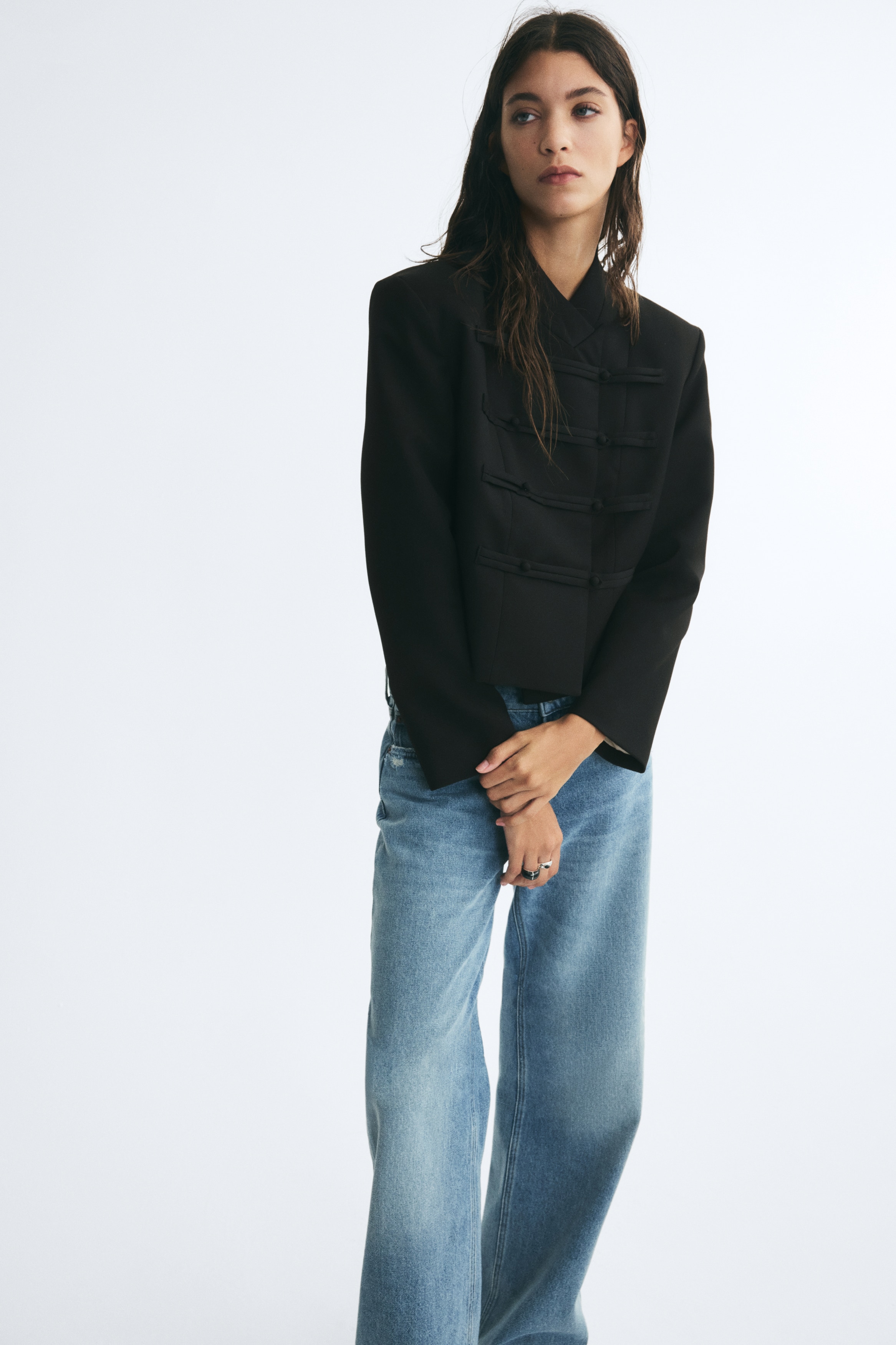 A black cropped jacket is shown facing front, featuring a structured, high wrap-over collar and long, straight sleeves. The front displays four horizontal rows of fabric-covered toggle loop closures.
