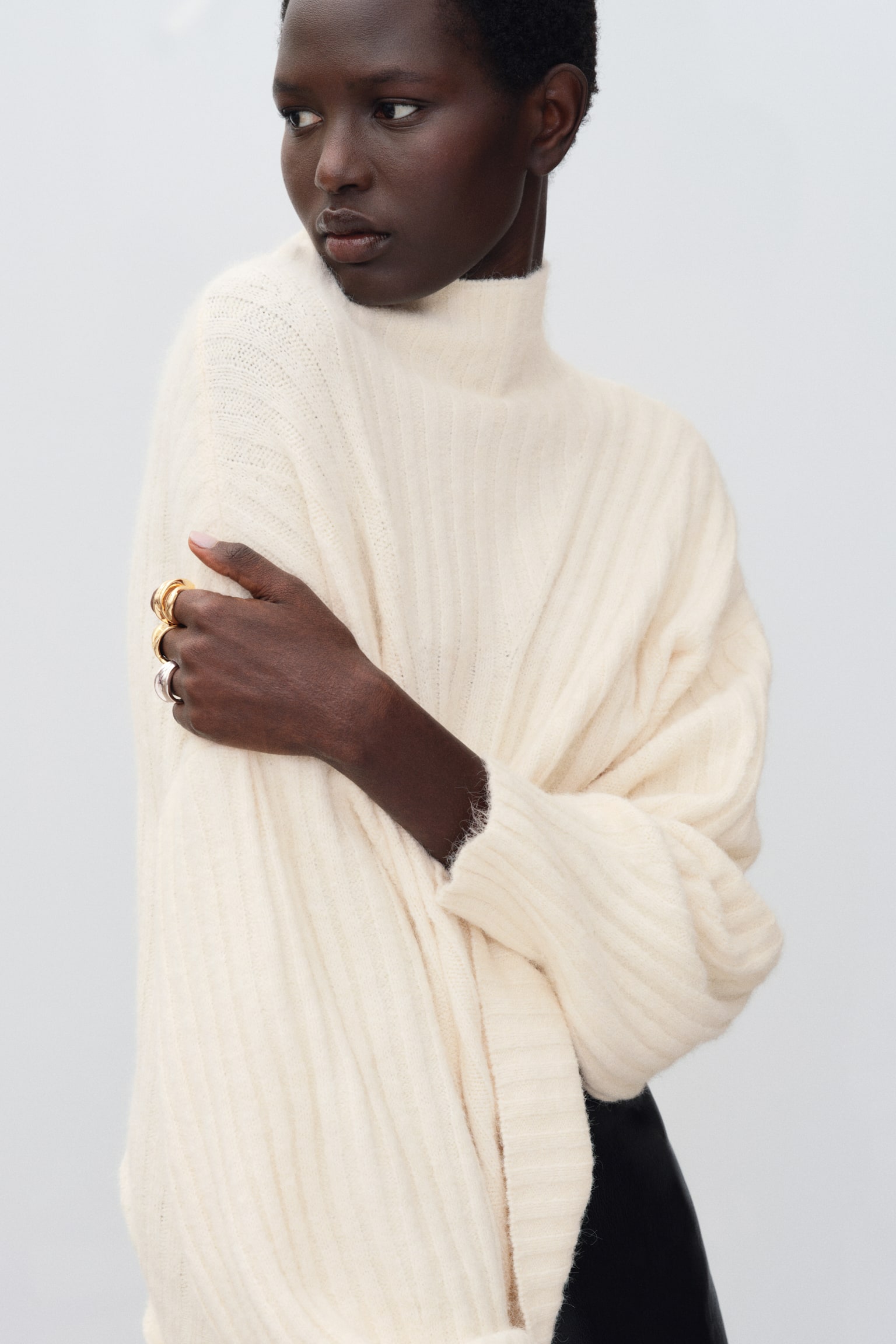 The model is wearing an off-white rib-knit sweater featuring a high neck and dropped shoulders. With one arm crossed over the chest to hold the opposite sleeve, the pose highlights the fabricâs soft texture. - 1