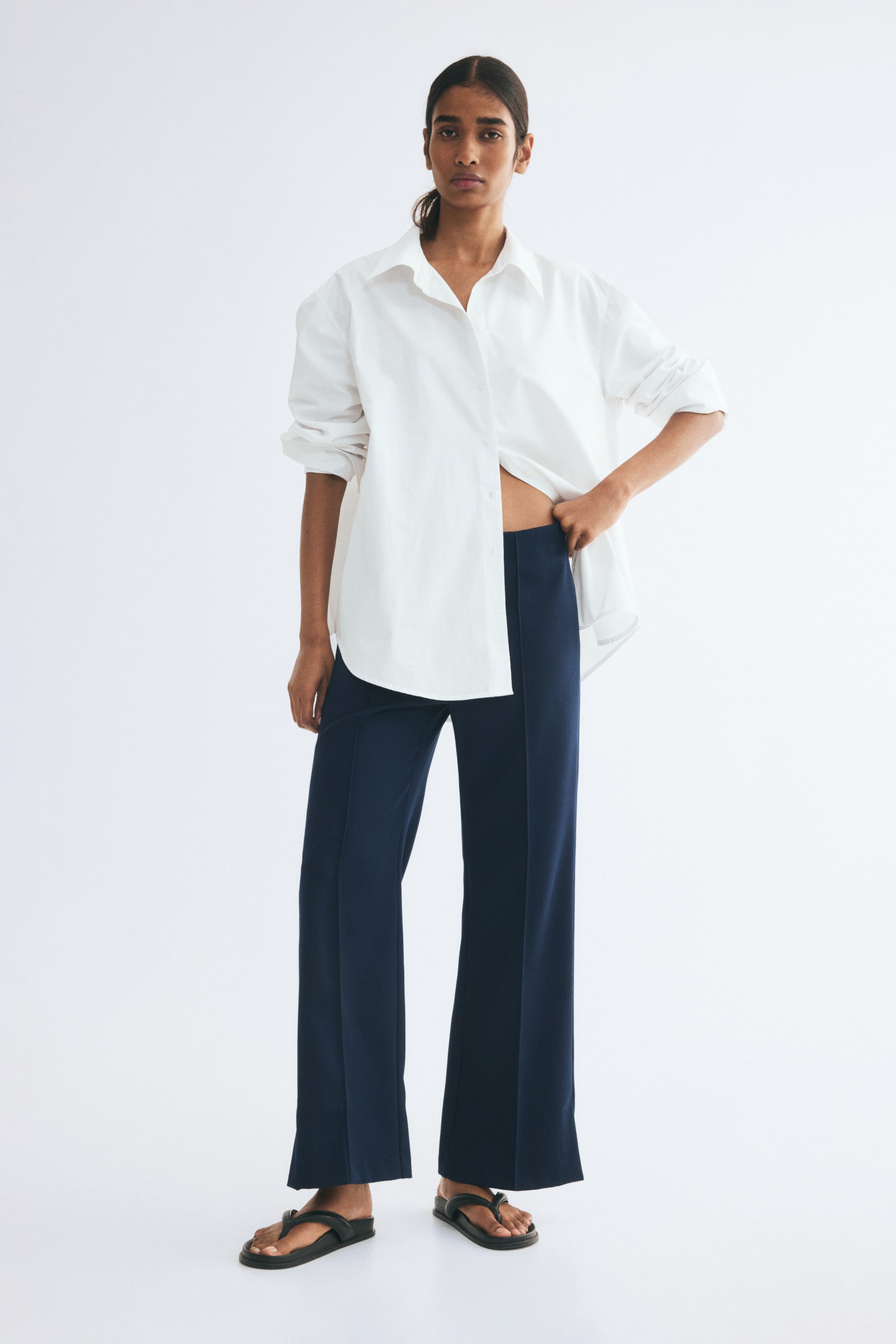 View larger image: The model is wearing dark blue wide-leg trousers with front creases. The trousers are styled with an oversized white button-down shirt with rolled-up sleeves and black flat sandals.