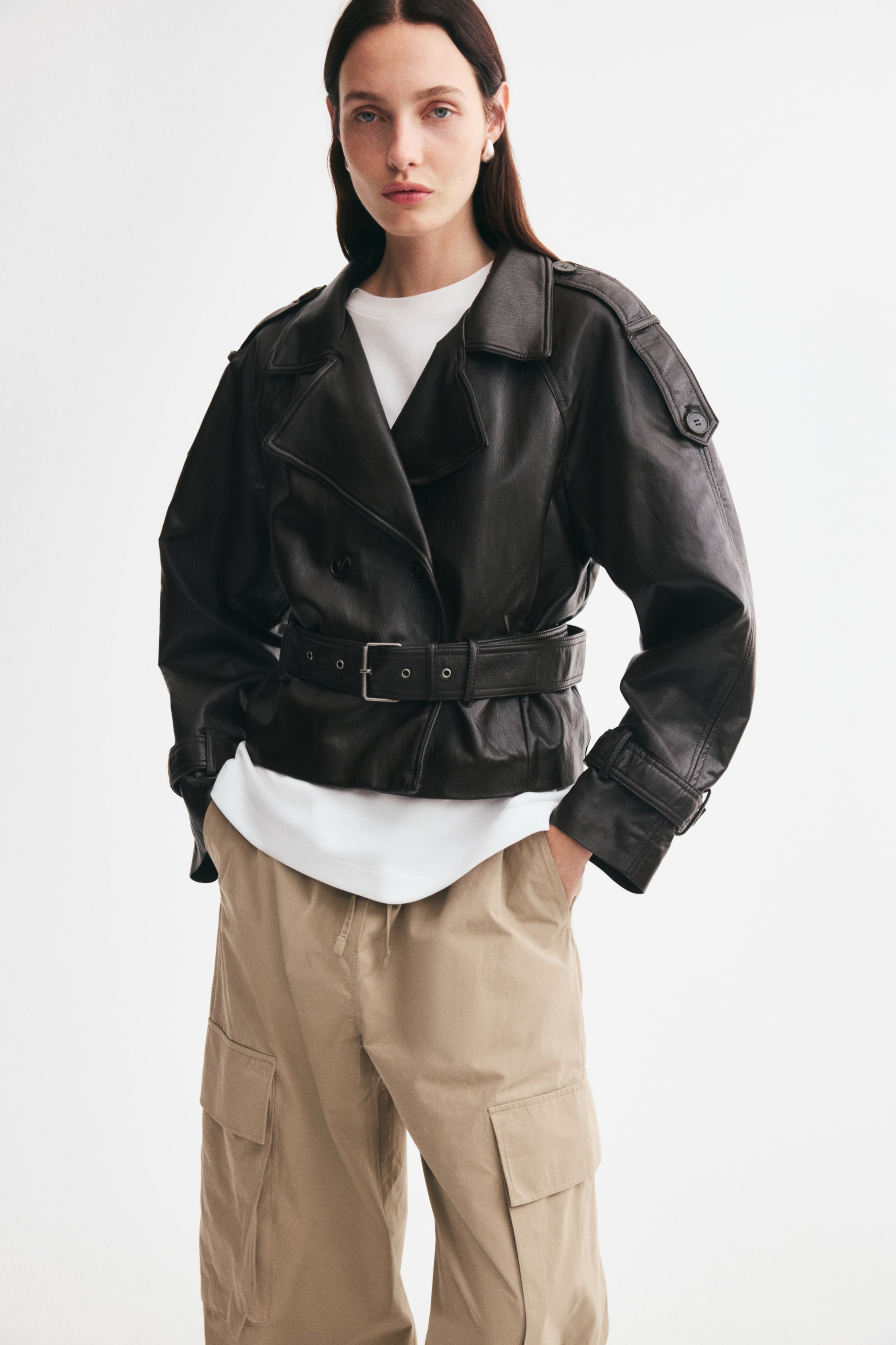 View larger image: The model is wearing a short black double-breasted biker jacket in coated fabric, featuring wide notch lapels, buttoned shoulder tabs, and an adjustable belt with a metal buckle fastened at the waist. The jacket is layered over a white T-shirt and paired with beige cargo pants.
