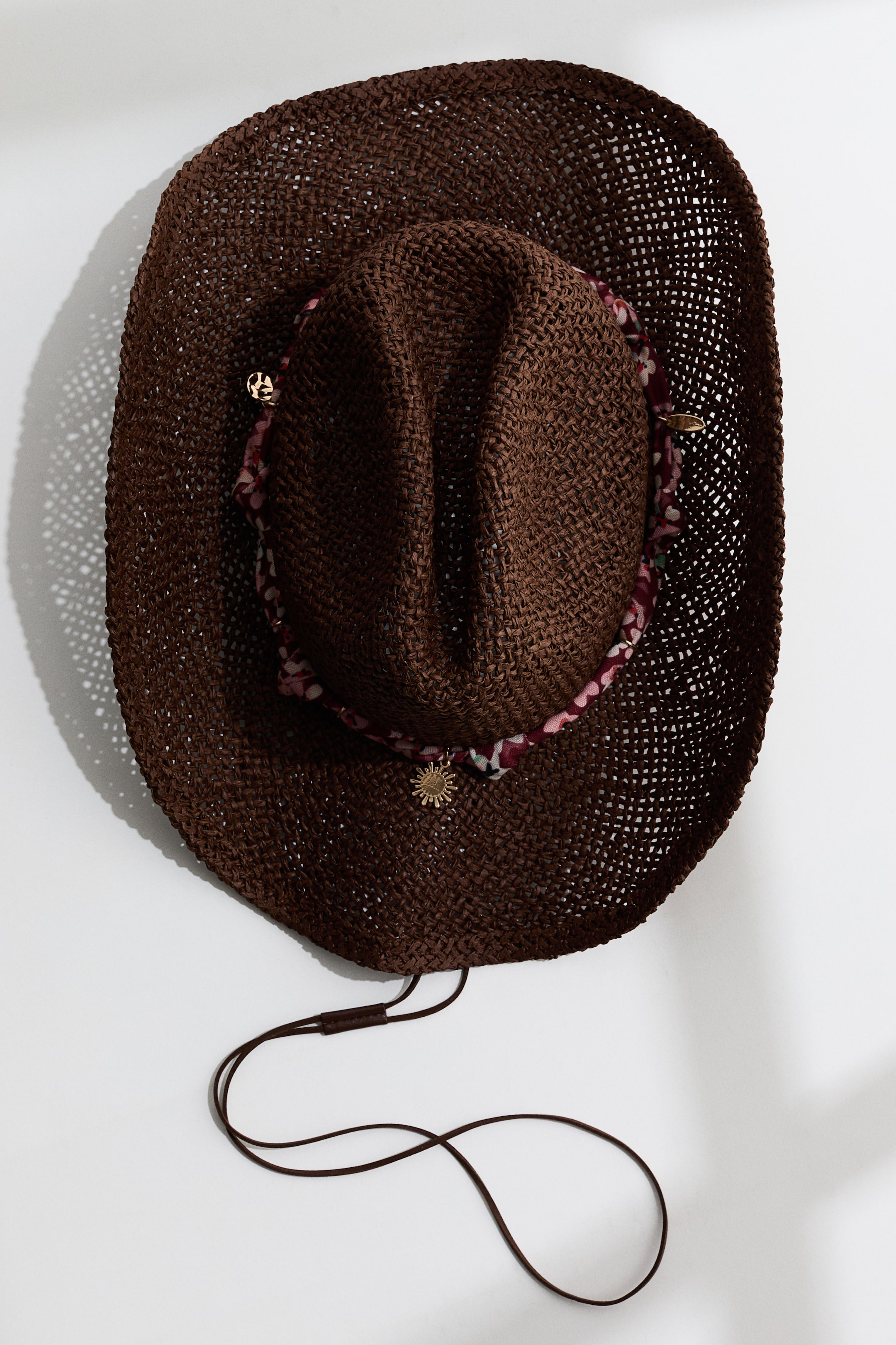 View larger image: A dark brown woven cowboy hat is lying flat, showing a top-down view of its creased crown and wide brim. A patterned fabric band, decorated with small gold-toned charms, encircles the base of the crown, and a thin brown cord chinstrap extends from the hat.