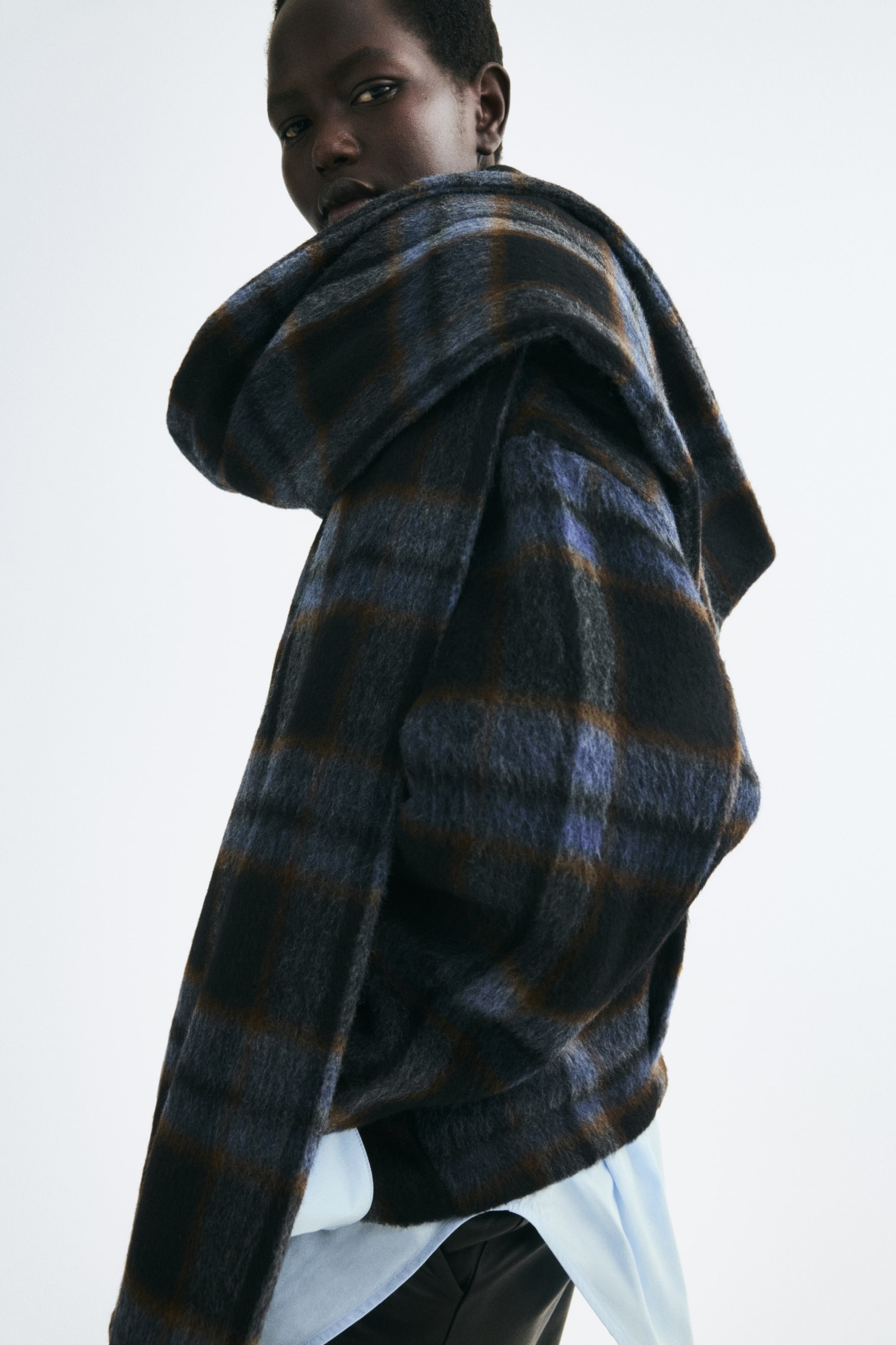 Voir une image plus grande: Une veste à carreaux surdimensionnée en tissu brossé bleu, brun et noir est portée par le mannequin, avec une écharpe intégrée assortie drapée sur l’épaule. La veste est superposée sur une chemise bleu pâle et un pantalon foncé.