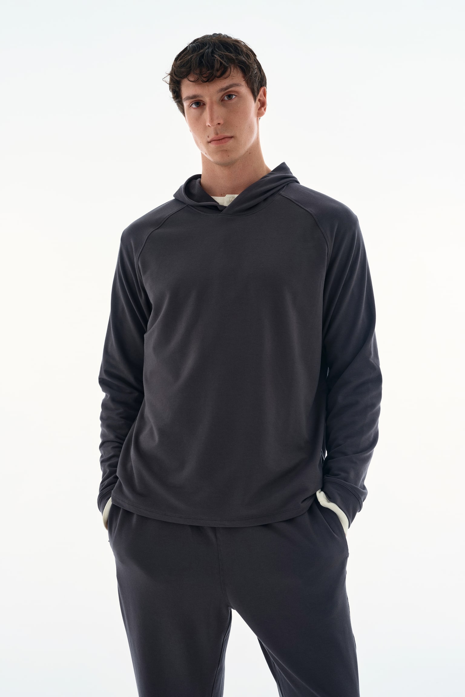 The model is wearing a dark grey long-sleeved hoodie, featuring a relaxed fit and raglan sleeves. The hoodie is layered over a cream-coloured top, visible at the collar and cuffs, and paired with matching dark grey sweatpants. - 1
