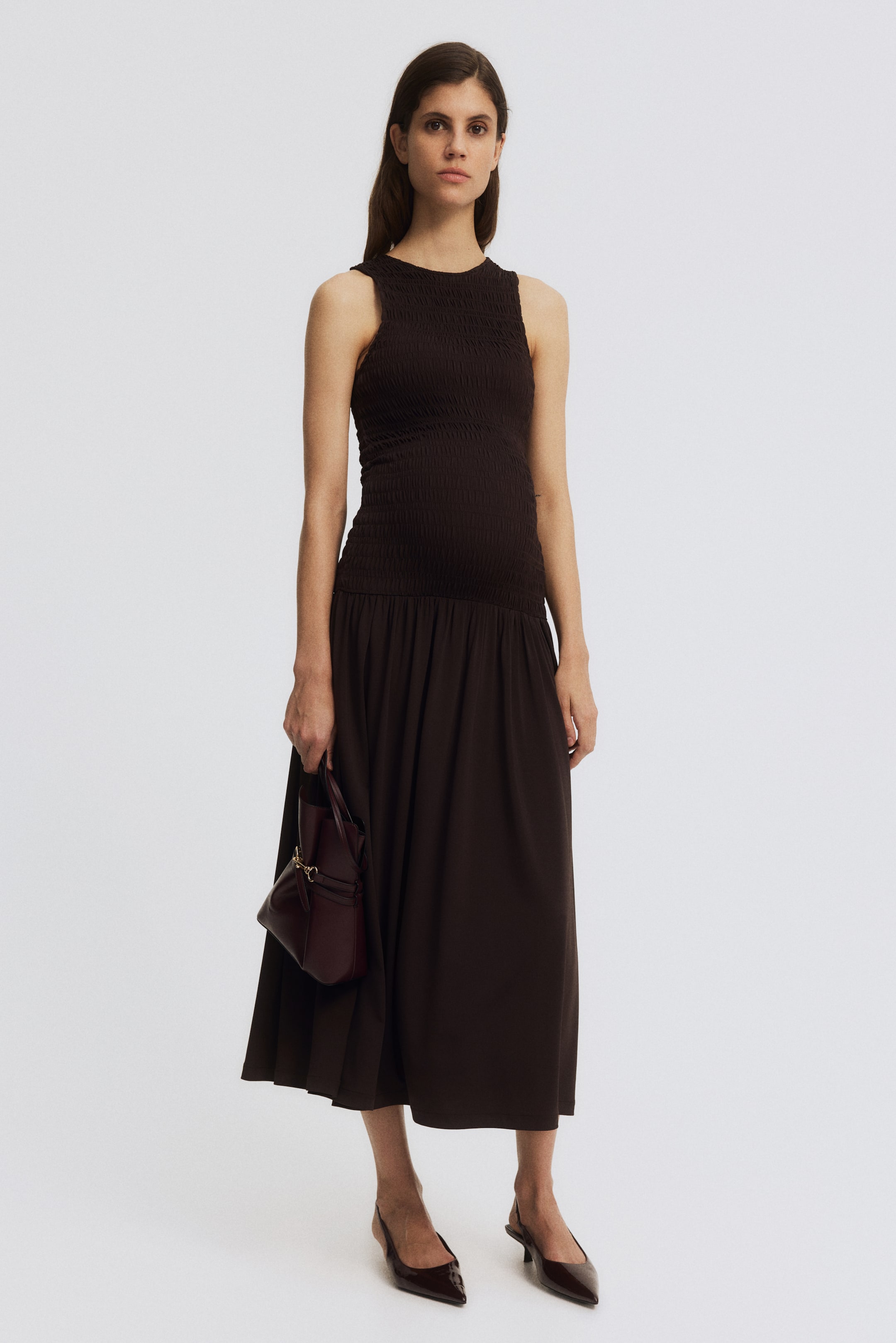 View larger image: A dark brown sleeveless dress features a fitted, shirred bodice with a round neck. It transitions into a flowing midi skirt that drapes to the ankles, showcasing its full front view.