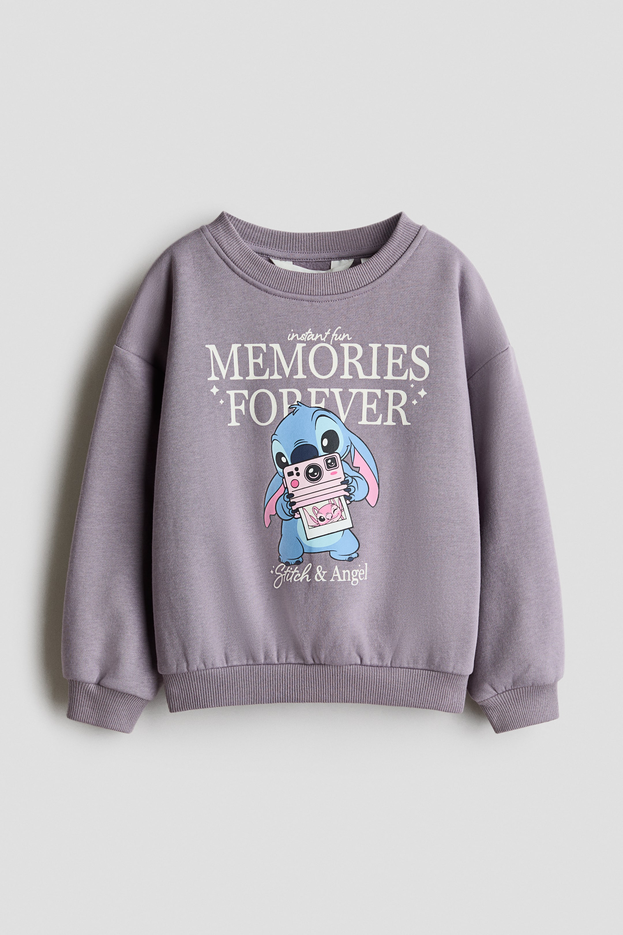 View larger image: A front-facing purple top in sweatshirt fabric features a round ribbed neckline, dropped shoulders, and long sleeves with ribbed cuffs and hem. The front showcases a graphic print of the text "instant fun MEMORIES FOREVER" above a blue character holding a pink instant camera, with "Stitch & Angel" printed below.