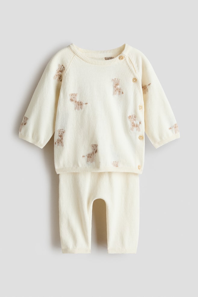 A cream-colored, fine-knit set, comprising a cardigan and trousers, is showing from the front, both pieces featuring a scattered pattern of light brown dog figures. The cardigan is displaying a round, rib-trimmed neckline with a side opening secured by three small buttons, along with ribbed cuffs and hem, while the trousers are presenting an elasticated, rib-trimmed waist and ribbed hems. - 1