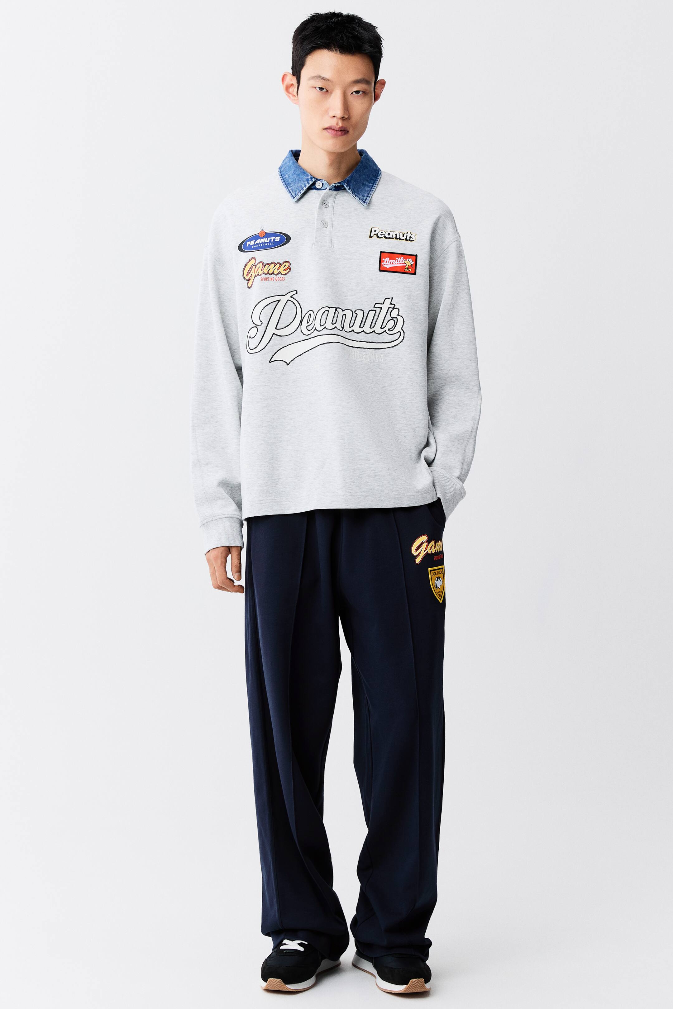 View larger image: The model is wearing a light grey long-sleeved polo sweatshirt featuring various "Peanuts" graphic patches and script. The sweatshirt is styled over a blue denim collar and paired with wide-leg navy blue trousers.