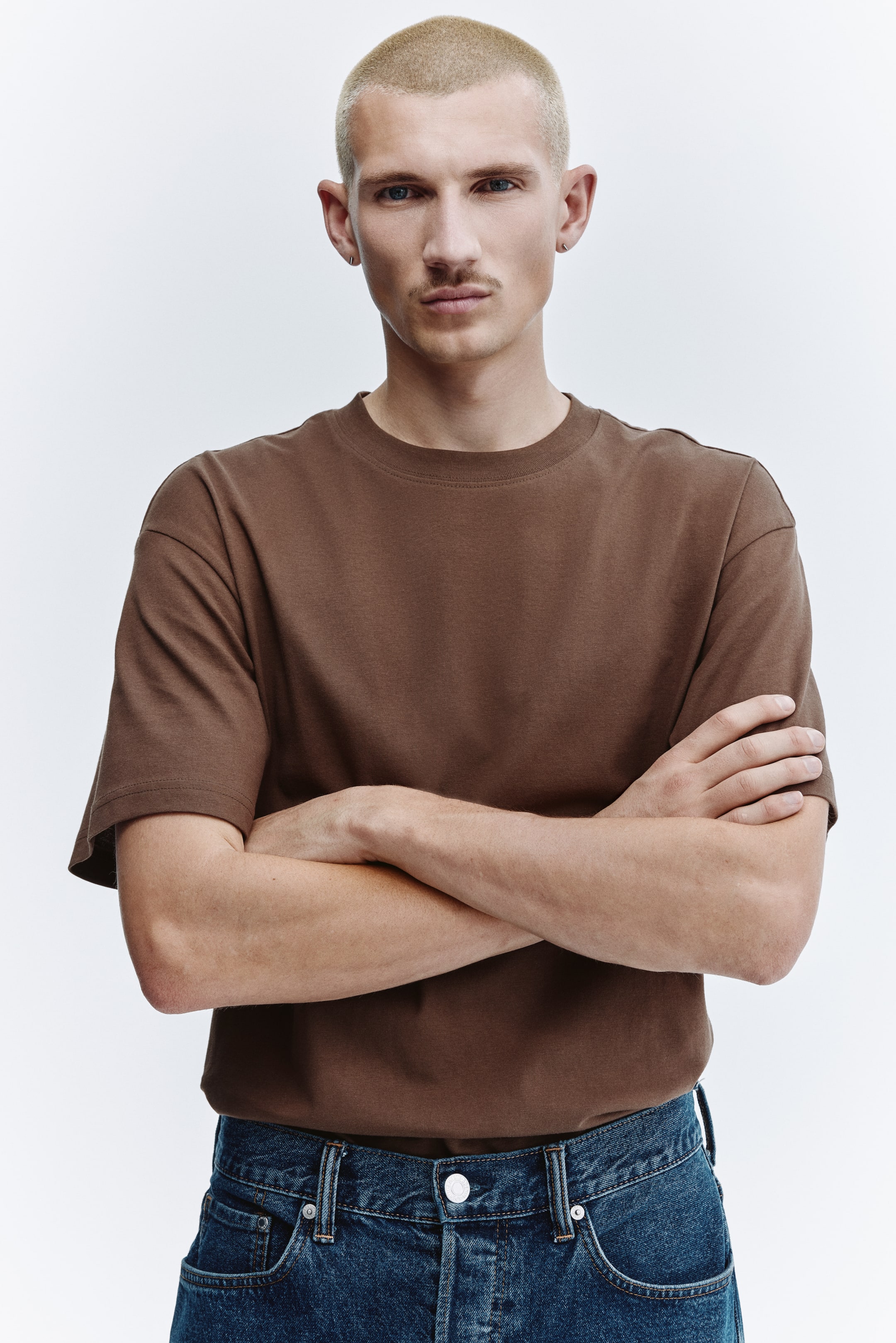 View larger image: A brown T-shirt features a crew neck and relaxed short sleeves. It offers a loose fit and is shown tucked into jeans.
