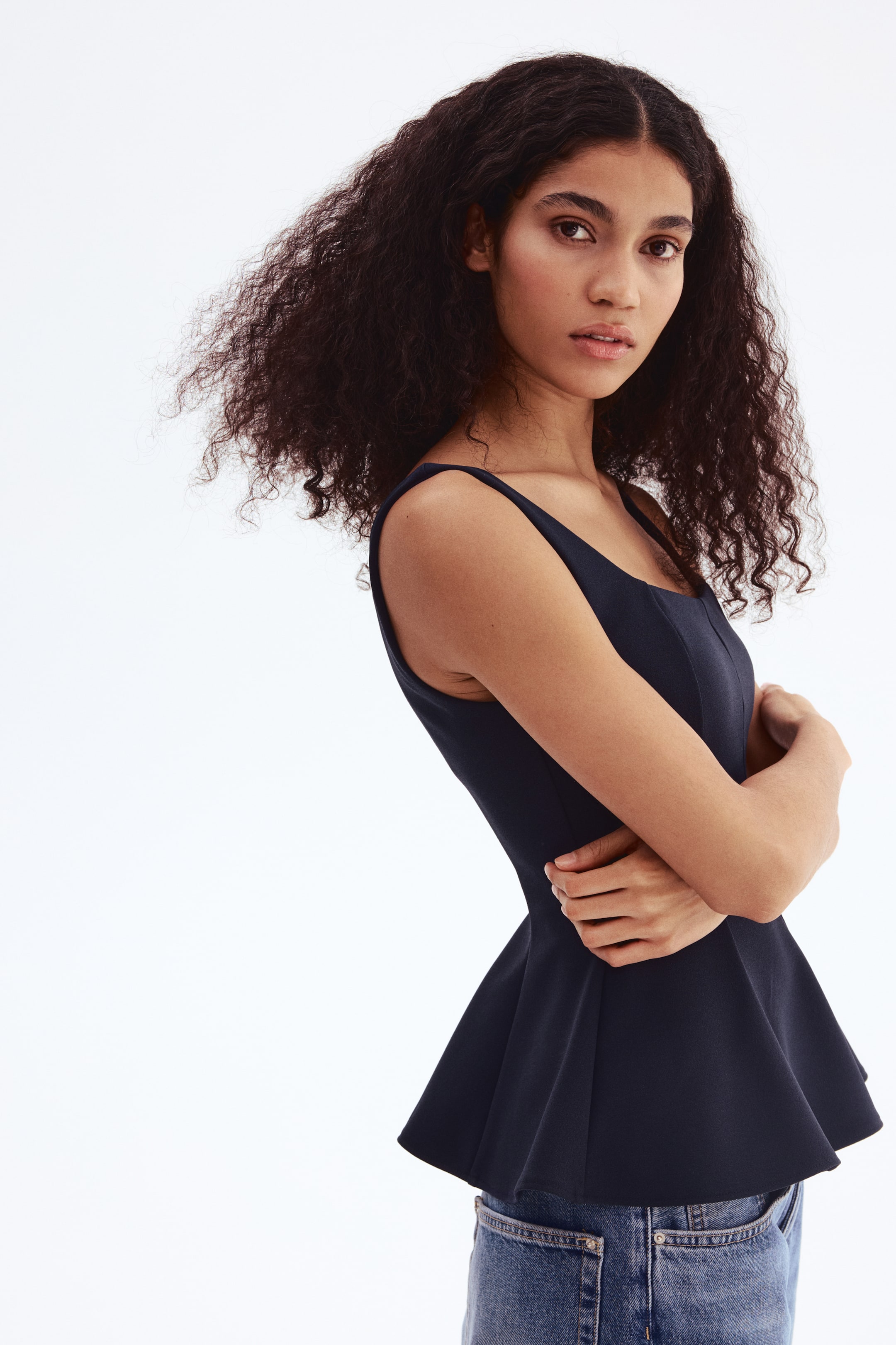 View larger image: A dark sleeveless peplum top features a square neckline and a fitted bodice that flares into a peplum hem. The top is shown from a slight side-front angle, resting above blue jeans.