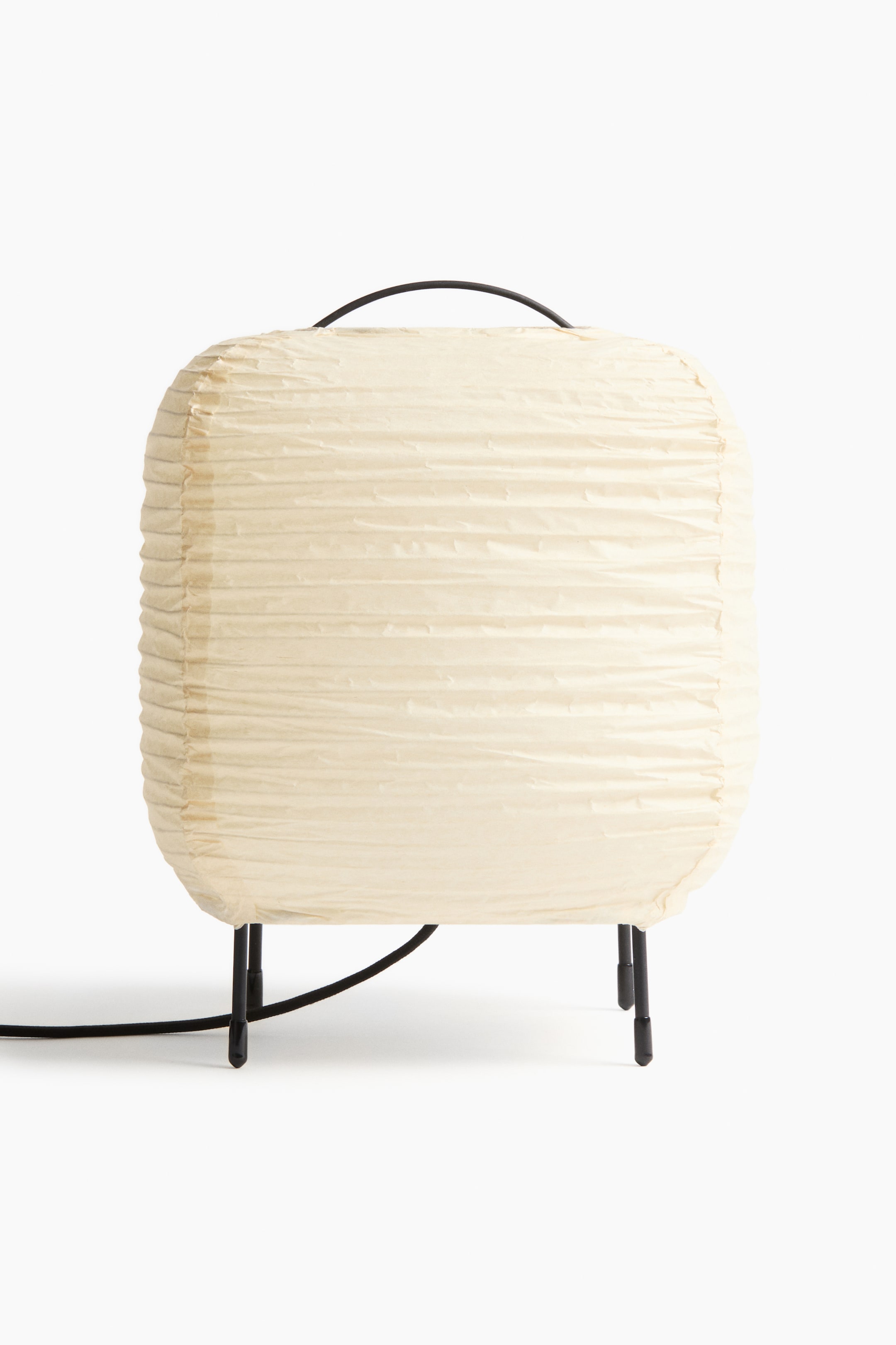 View larger image: A rectangular lamp featuring a light cream, horizontally pleated paper shade. It stands on a black metal base with four slender legs, and a black arched handle rises from its top, with an electrical cord extending from the side.