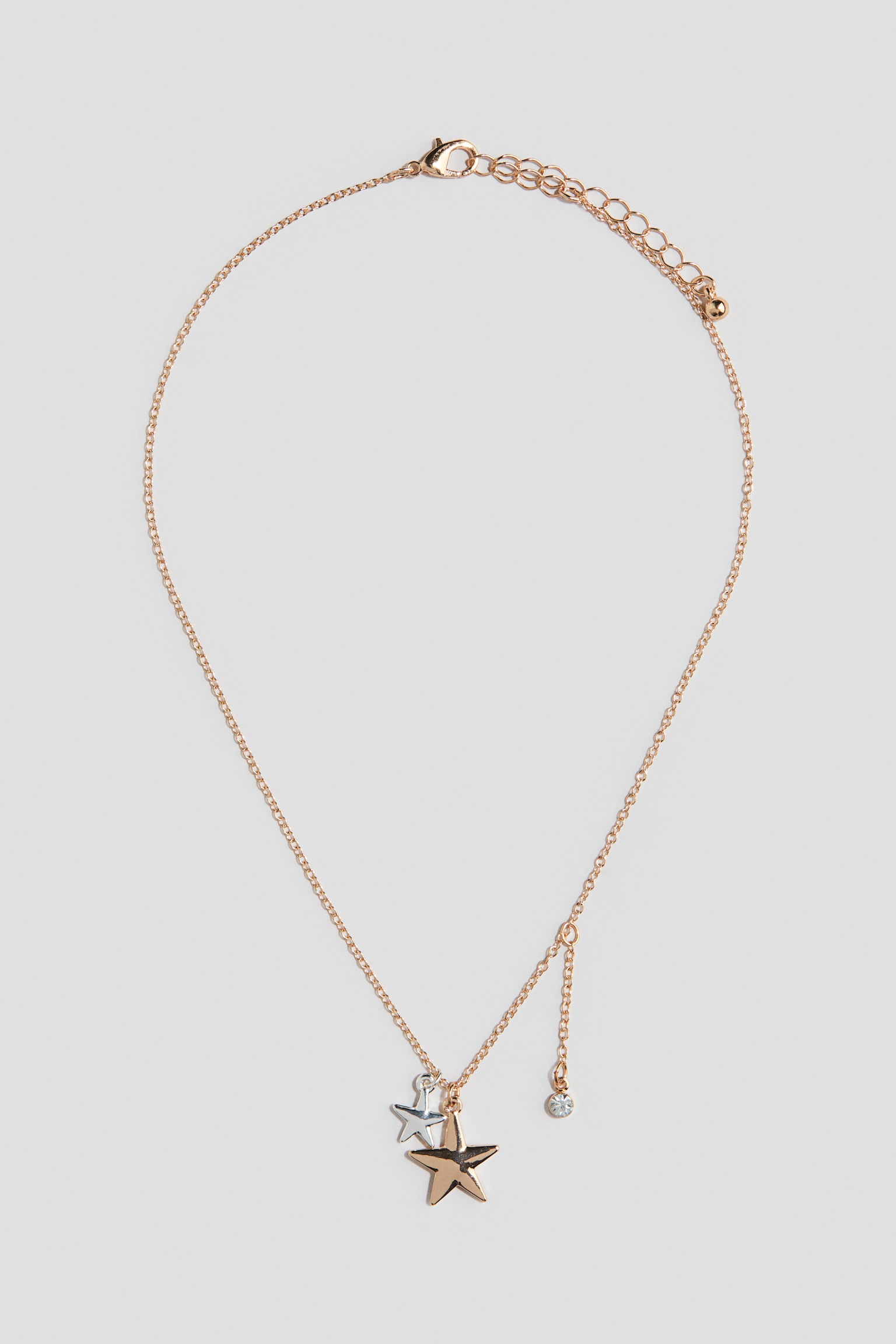 Eine feine Kette in Goldton liegt flach in einer Vorderansicht und zeigt einen grÃ¶sseren SternanhÃ¤nger in Goldton, der mit einem kleineren SternanhÃ¤nger in Silberton in der Mitte geschichtet ist. An der Hauptkette hÃ¤ngt seitlich eine kurze, zarte Kette herab, die in einem kleinen, runden StrassanhÃ¤nger endet.