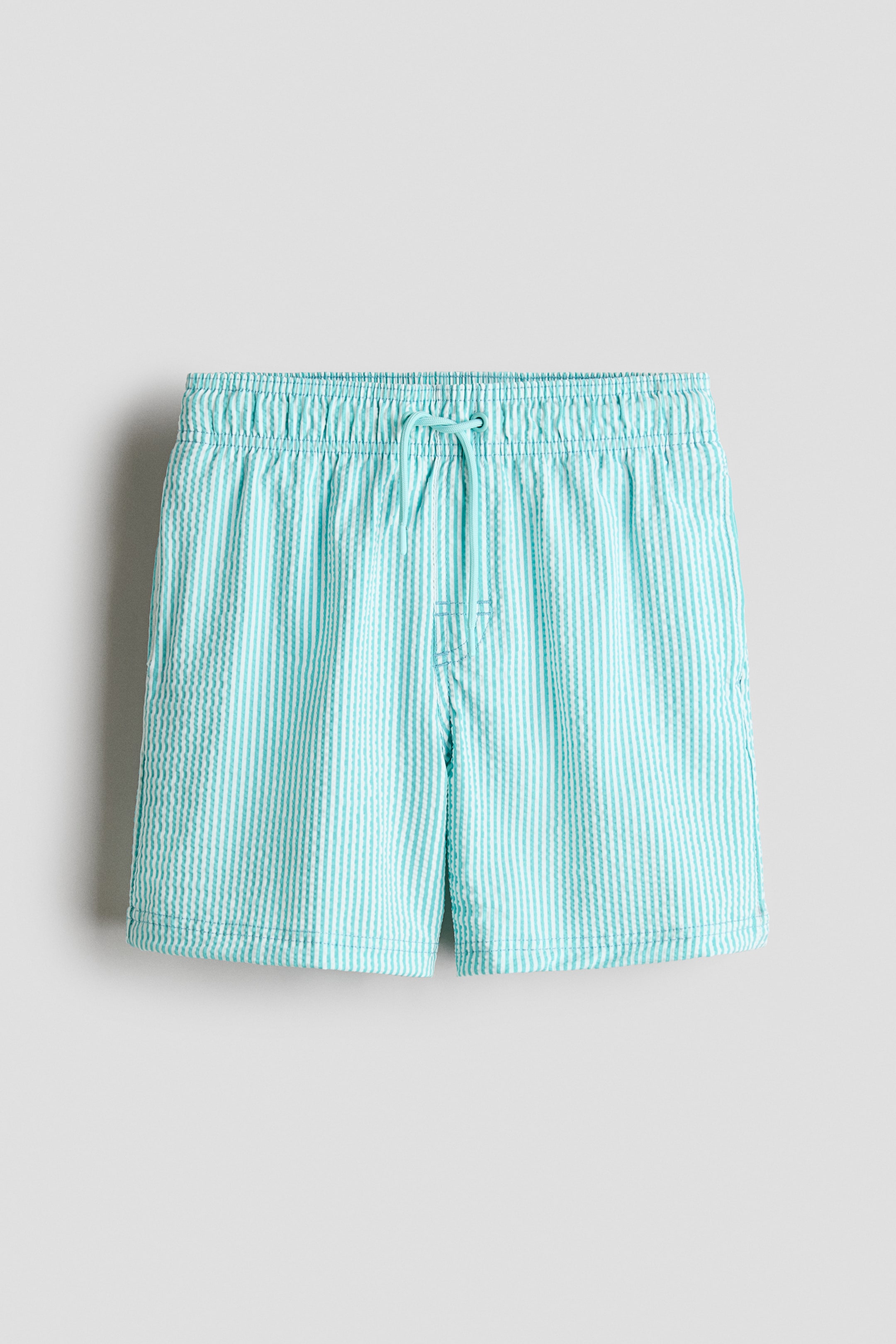 Größeres Bild anzeigen: Badeshorts werden von vorne präsentiert und zeigen hellblaue und weiße vertikale Seersucker-Streifen. Sie sind mit einem elastischen Bund, einer sichtbaren Kordel, einem Fake-Hosenschlitz und dezenten Seitentaschen gestaltet.