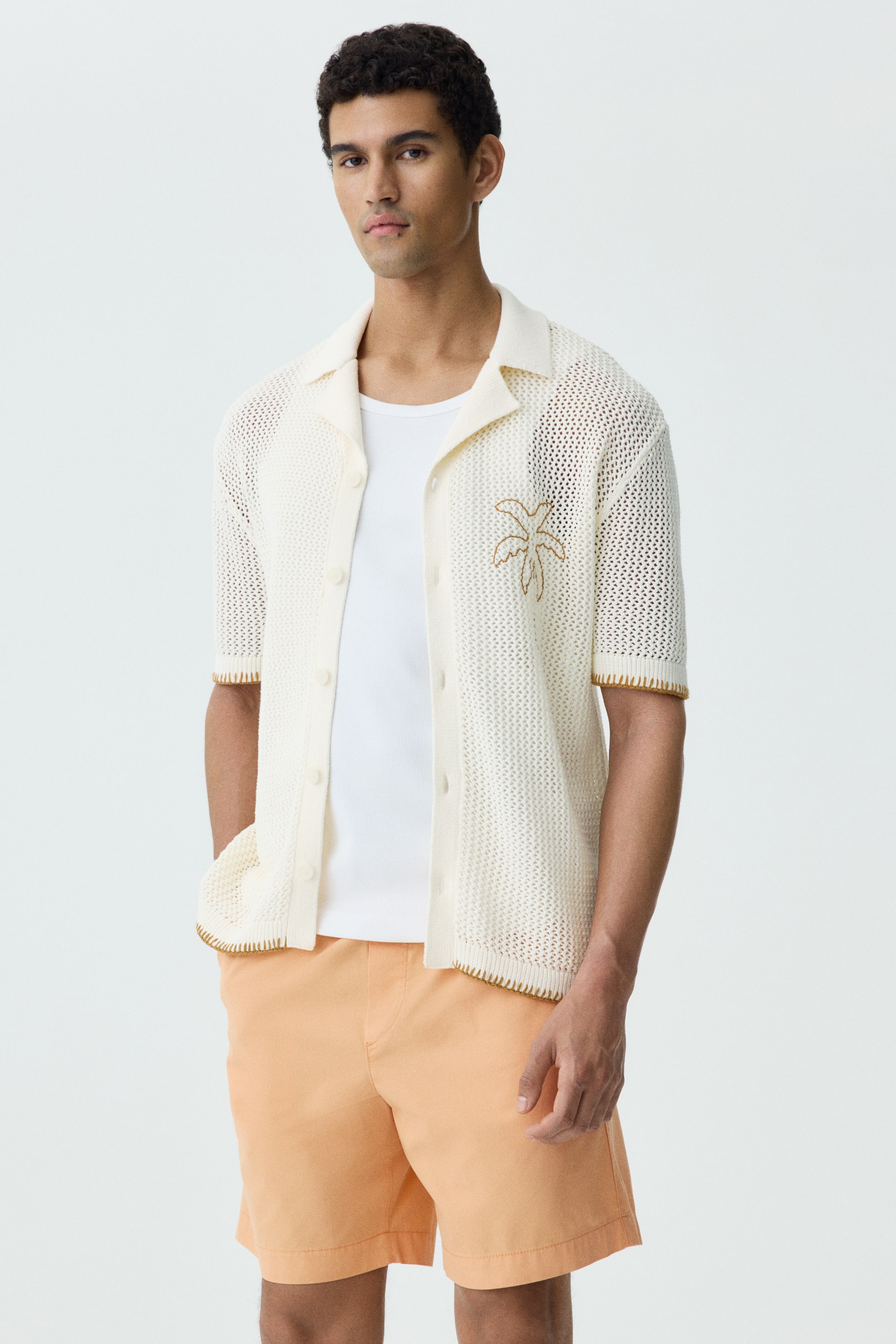 View larger image: A cream open-knit short-sleeved shirt with a camp collar and button-front closure is styled open over a white t-shirt, showcasing a brown embroidered palm tree on the left chest. It features contrasting brown stitching along the collar, placket, cuffs, and hem, and is paired with light orange flat-front shorts.