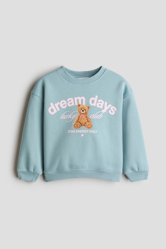 A light blue sweatshirt is shown from the front, featuring a round ribbed neckline, dropped shoulders, and ribbed cuffs and hem. The front features a printed design of a brown teddy bear, surrounded by pink text that reads "dream days", "lucky club", and "STAR ENERGY ONLY." - 1