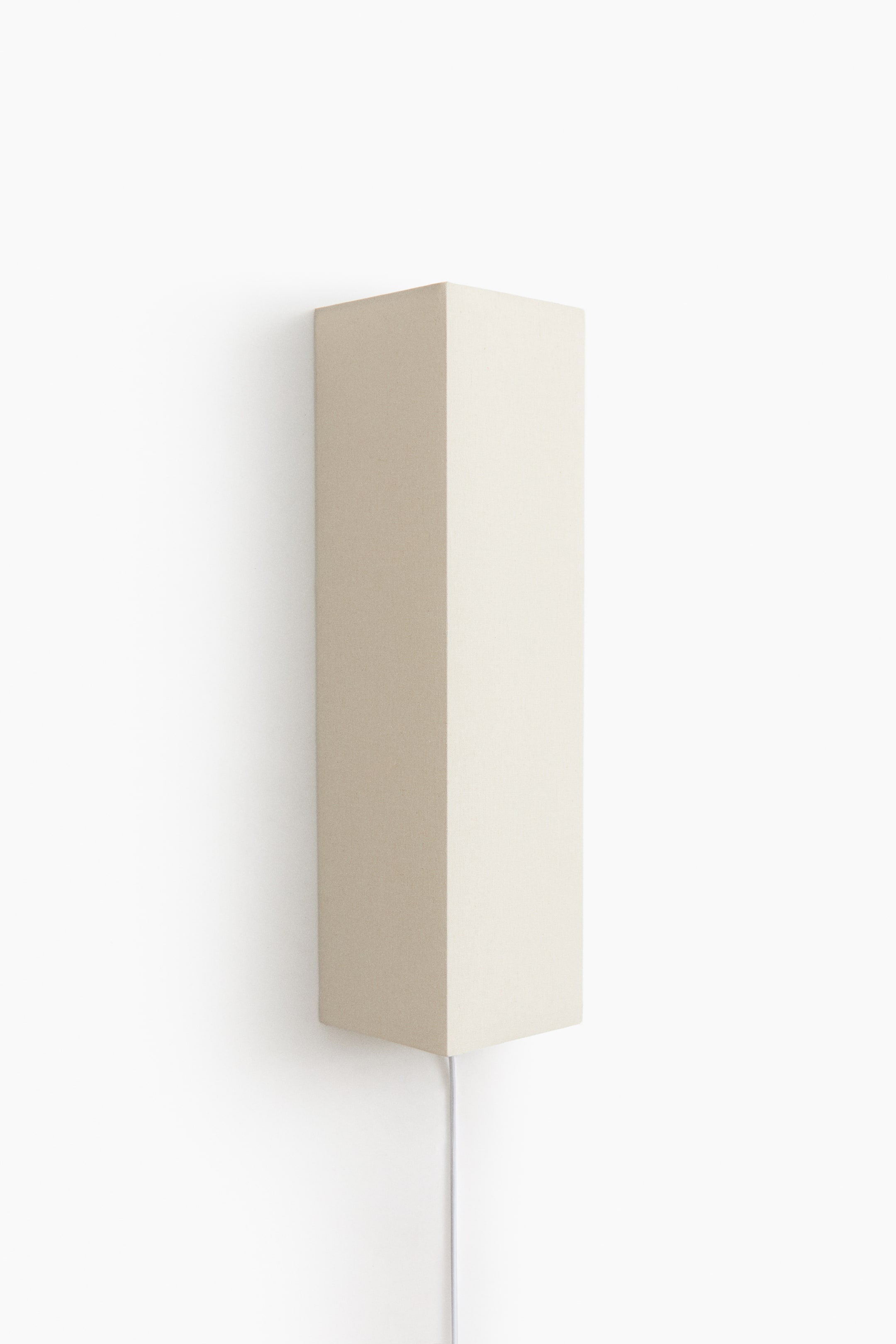 View larger image: A wall-mounted sconce lamp, shown from a front angle, features a tall, rectangular column shade covered in a textured, light beige fabric. A thin white electrical cord extends straight down from the bottom centre of the shade.