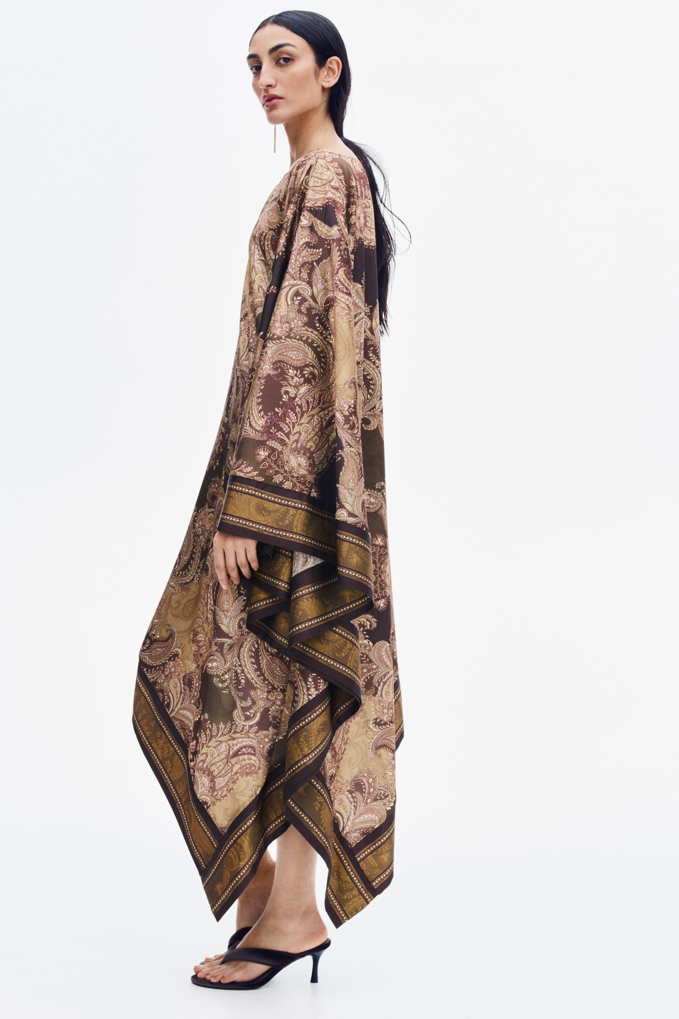 View larger image: A satin cape dress featuring a brown and olive green paisley print is being worn by the model. The dress has a loose, draped silhouette with an asymmetrical handkerchief hem and is paired with dark heeled thong sandals.