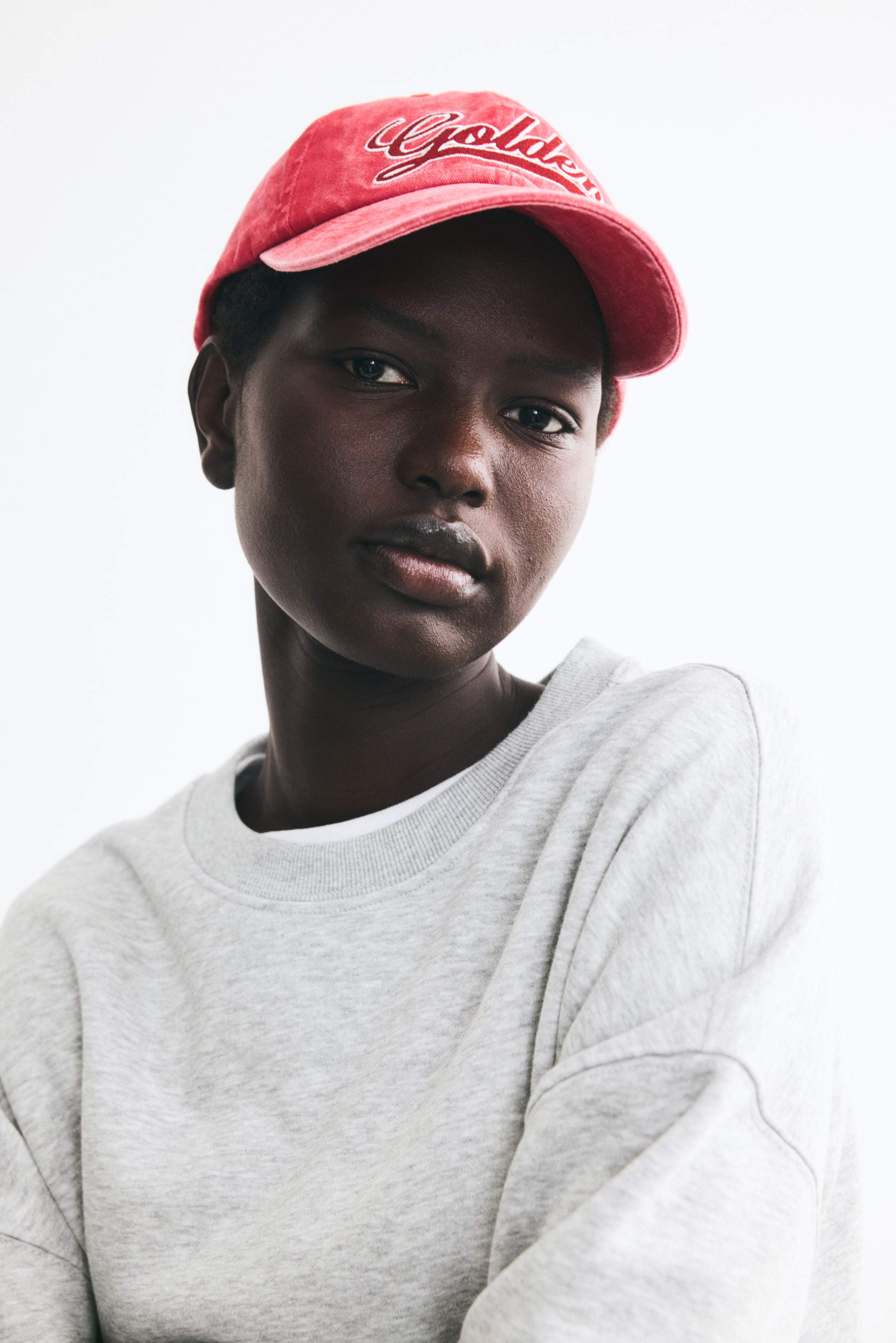 View larger image: A light grey melange oversized sweatshirt is being worn by the model, featuring a ribbed round neckline and dropped shoulders. The sweatshirt is layered over a white t-shirt, and the model is also wearing a red baseball cap.