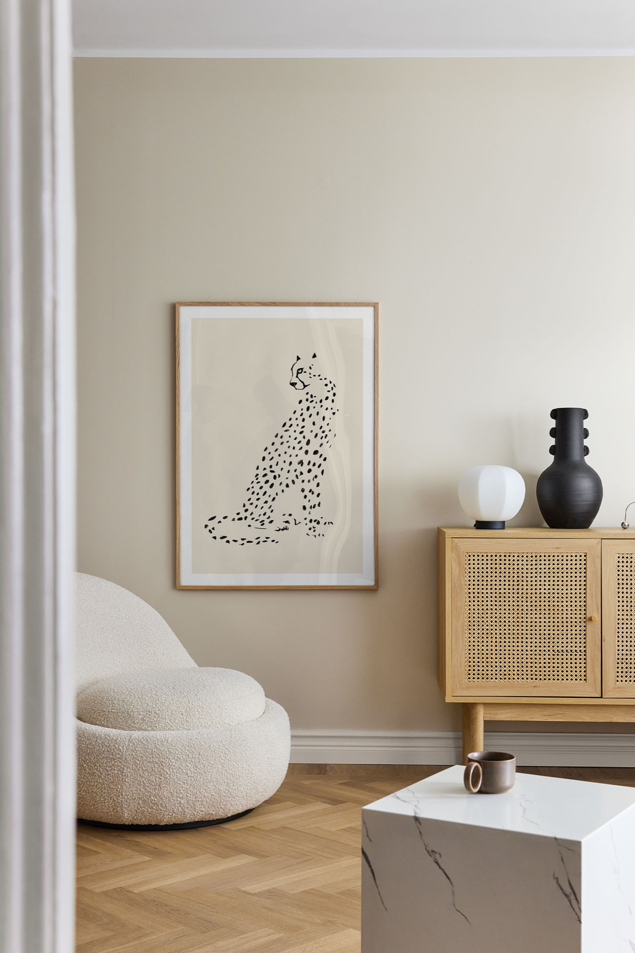 Größeres Bild anzeigen: Ein gerahmter Kunstdruck zeigt eine minimalistische Schwarz-Weiß-Illustration eines sitzenden Geparden im Profil vor einem hellbeigen Hintergrund, eingefasst in einen schmalen, hellen Holzrahmen.