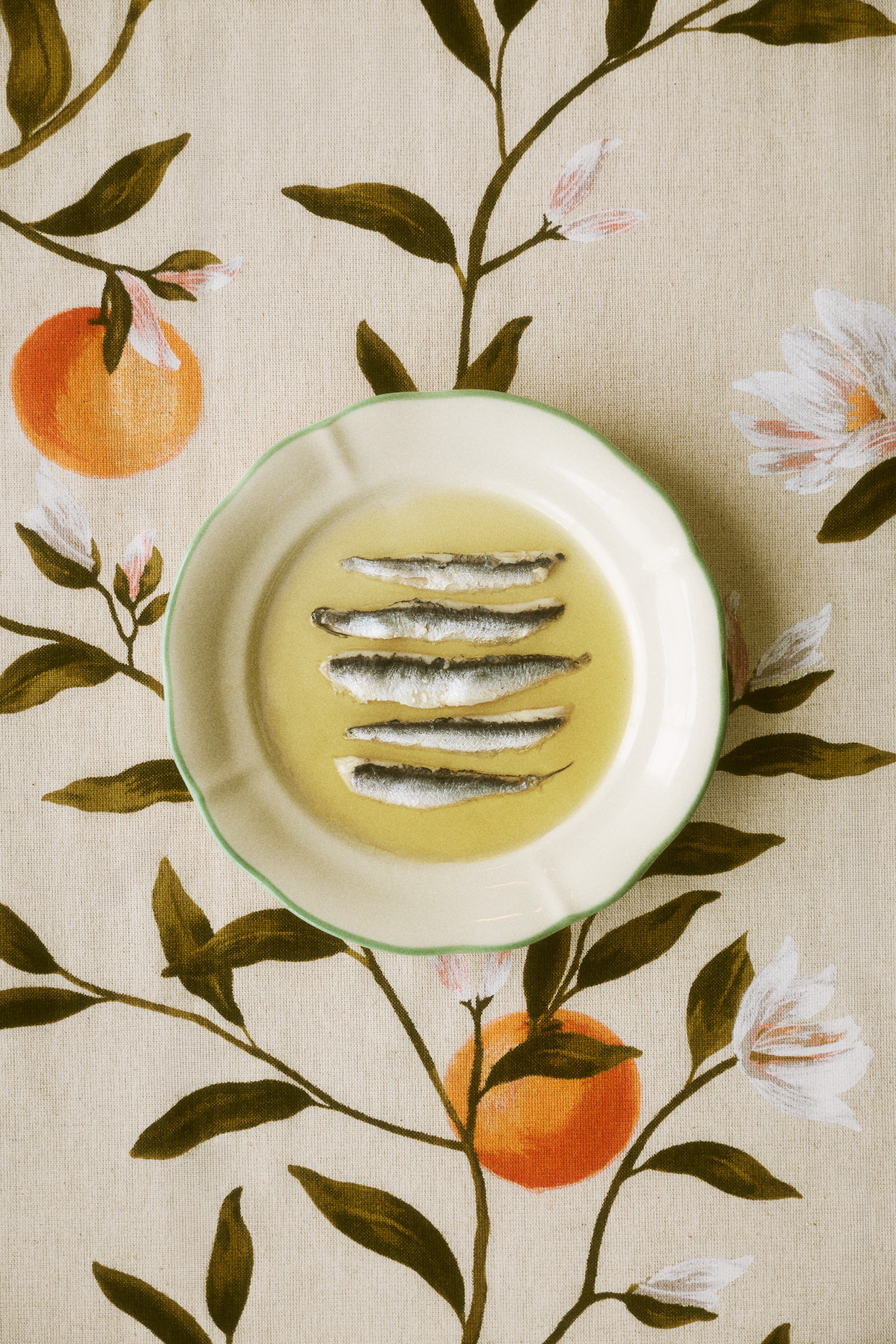 View larger image: Five sardines are arranged horizontally in clear yellow oil, resting on a round, white, fluted plate with a green rim, viewed from directly above.