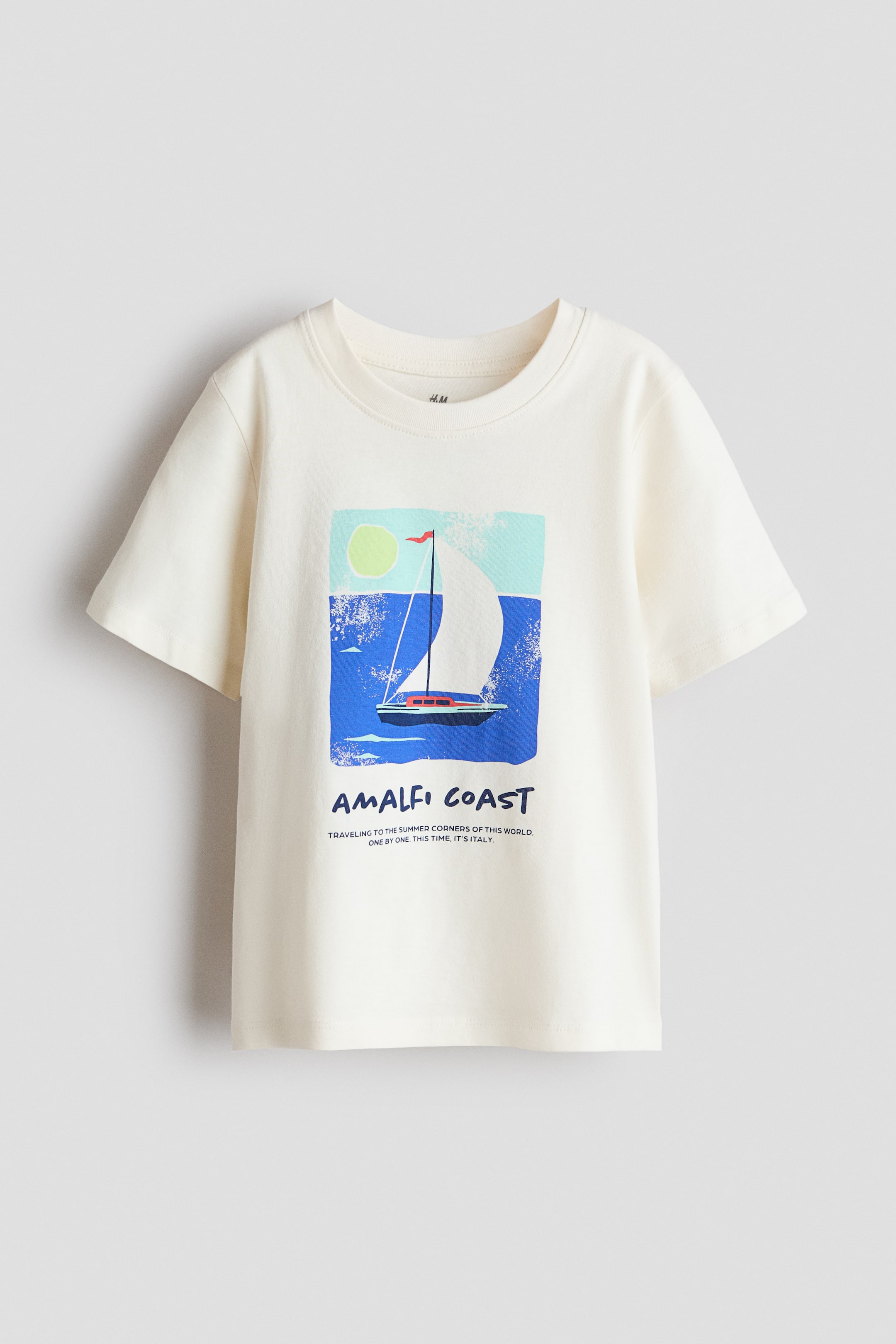 View larger image: A cream T-shirt is displayed as FRONT, featuring a ribbed crew neckline, dropped shoulders, and a straight-cut hem. Its front showcases a rectangular graphic print depicting a sailboat on blue water with a yellow sun in a light blue sky, above the text 'AMALFI COAST' and smaller text 'TRAVELING TO THE SUMMER CORNERS OF THIS WORLD. ONE BY ONE. THIS TIME, IT'S ITALY.'
