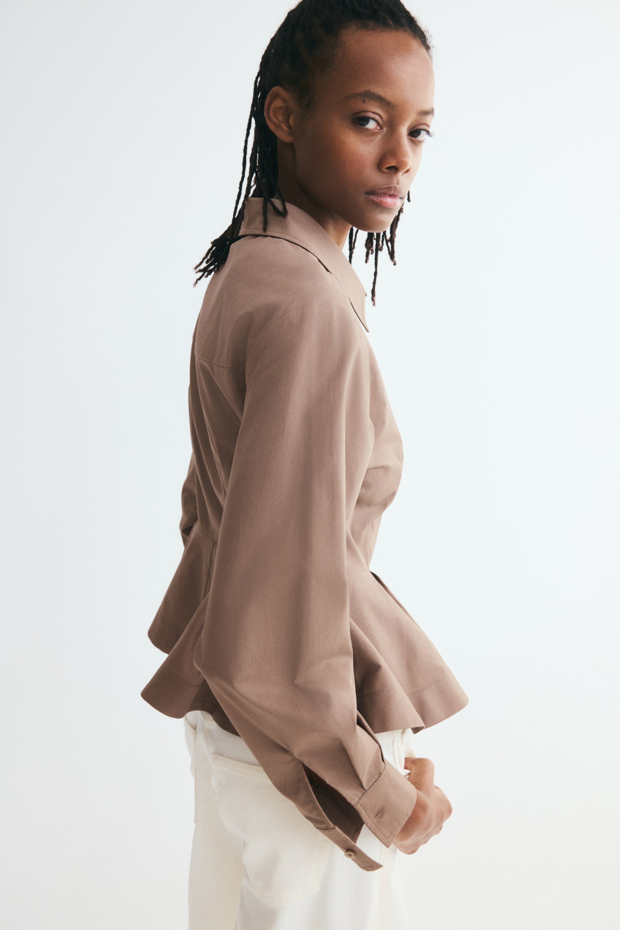 Visualizza l'immagine ingrandita: Una camicia in popeline di cotone taupe, rivolta verso destra, mette in risalto una silhouette strutturata con cuciture modellanti e un orlo peplum svasato e plissettato. La camicia presenta ampie maniche lunghe con polsini abbottonati ed è abbinata a pantaloni bianchi.