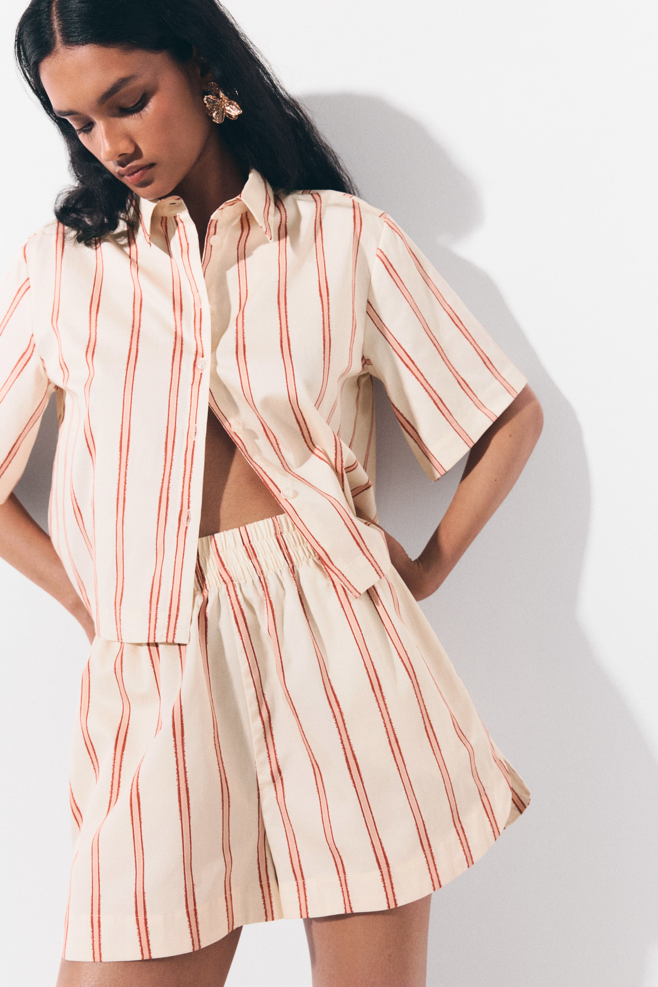 View larger image: This two-piece set, featuring a short-sleeved button-down shirt and matching shorts, is crafted from a light cream fabric with vertical red stripes. The collared shirt is worn open and cropped, while the coordinating shorts boast an elastic waistband and a loose fit.
