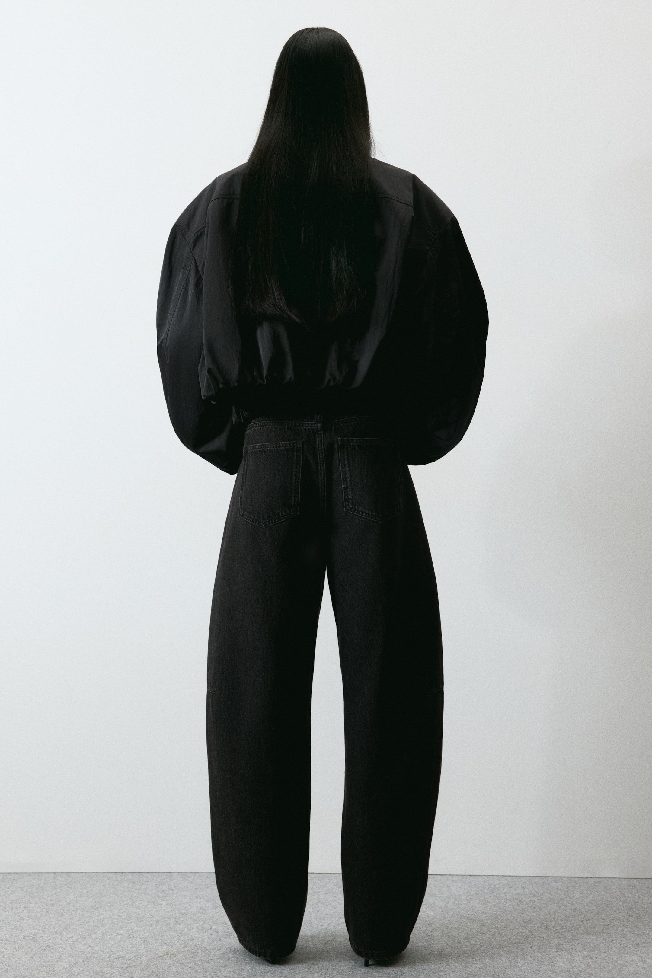 Ampliar la imagen: Una chaqueta bomber negra recortada y jeans negros de pierna ancha se presentan desde la vista trasera. La chaqueta bomber presenta mangas voluminosas y un dobladillo elástico, mientras que los jeans muestran un ajuste de talle alto con dos bolsillos traseros aplicados y una silueta de pierna ancha y redondeada.