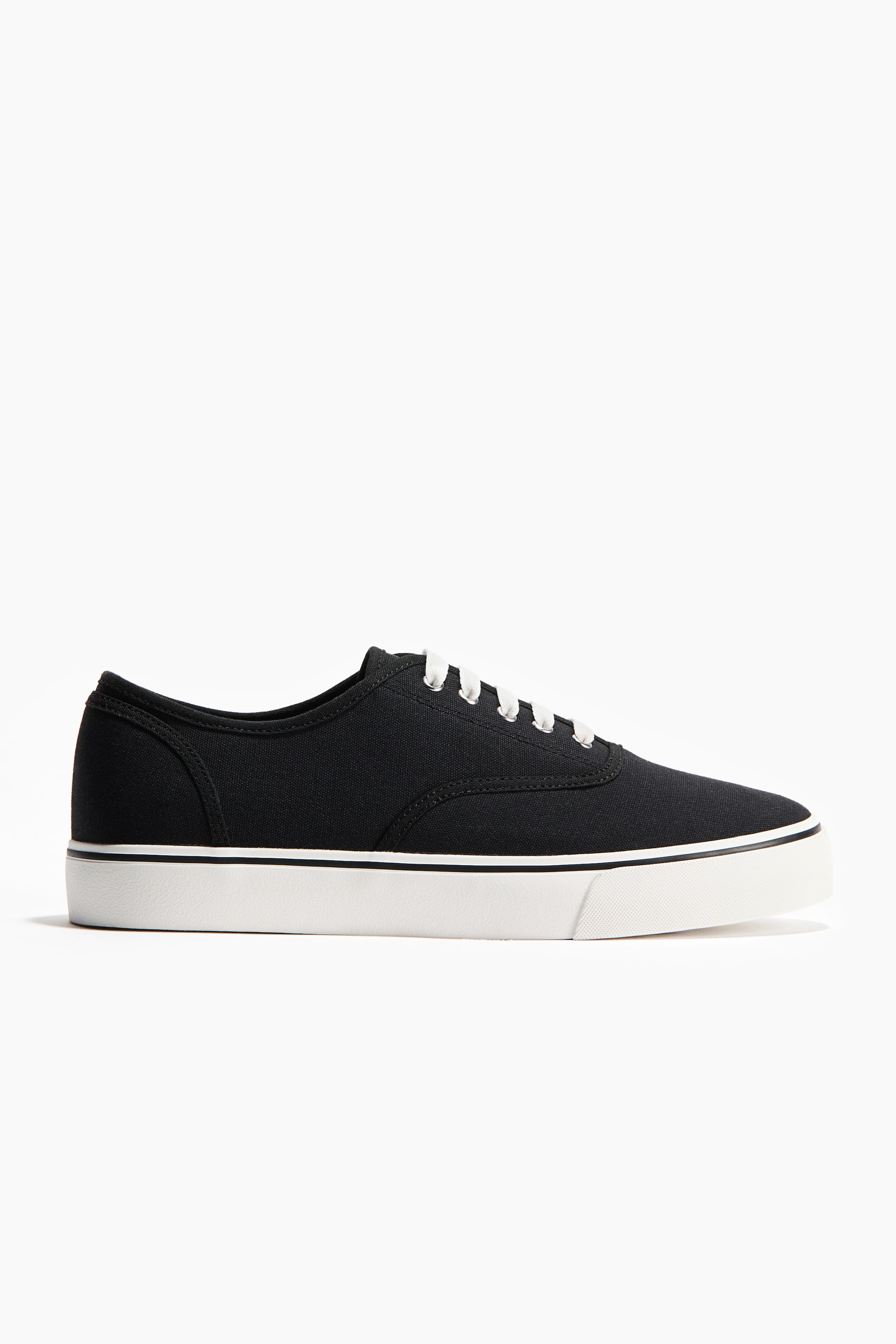 View larger image: A black canvas sneaker is shown in a side profile. It features white laces, a thick white sole, and a thin black stripe running along the sole's perimeter.