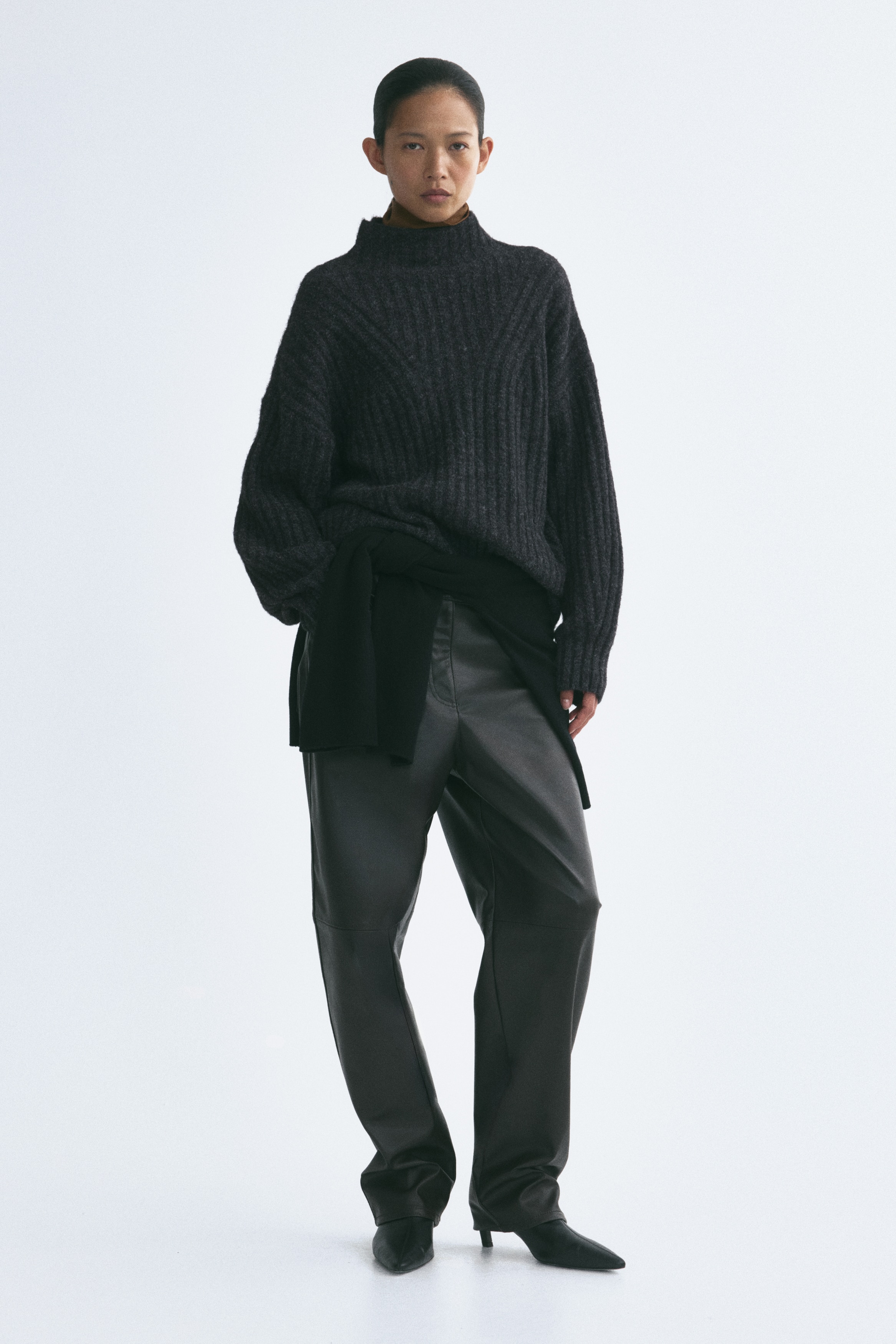 A dark grey jumper in a textured rib knit is shown from the front, featuring a mock turtleneck, dropped shoulders, and long sleeves with ribbed cuffs. The vertical ribbing pattern curves near the neckline and extends straight down to a straight hem.
