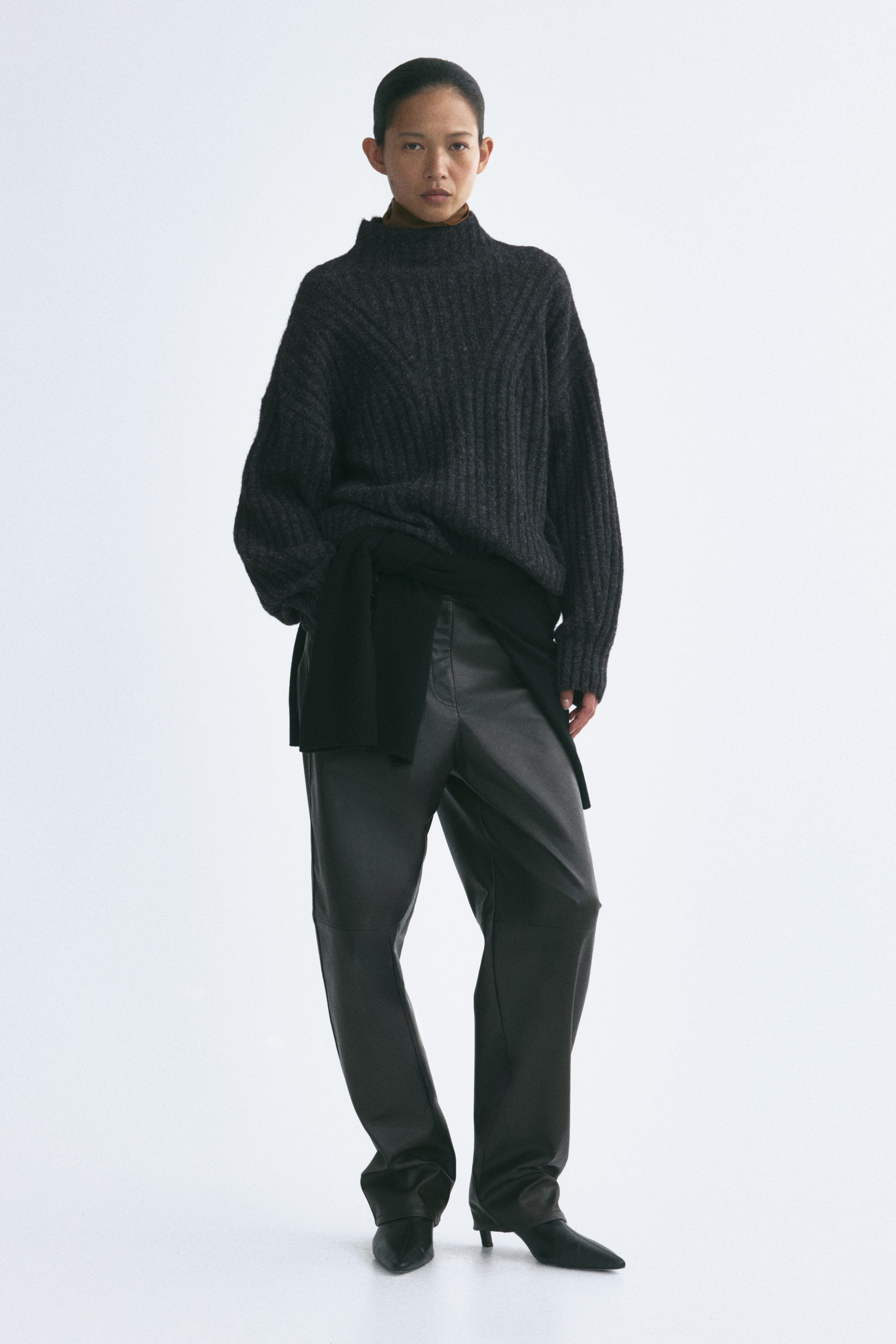 Agrandir l'image: Un pull en maille côtelée gris foncé, doté d’un col montant, de épaules tombantes et de manches longues, est porté par le mannequin. Le pull est associé à un vêtement noir noué autour de la taille et à un pantalon sombre ample.
