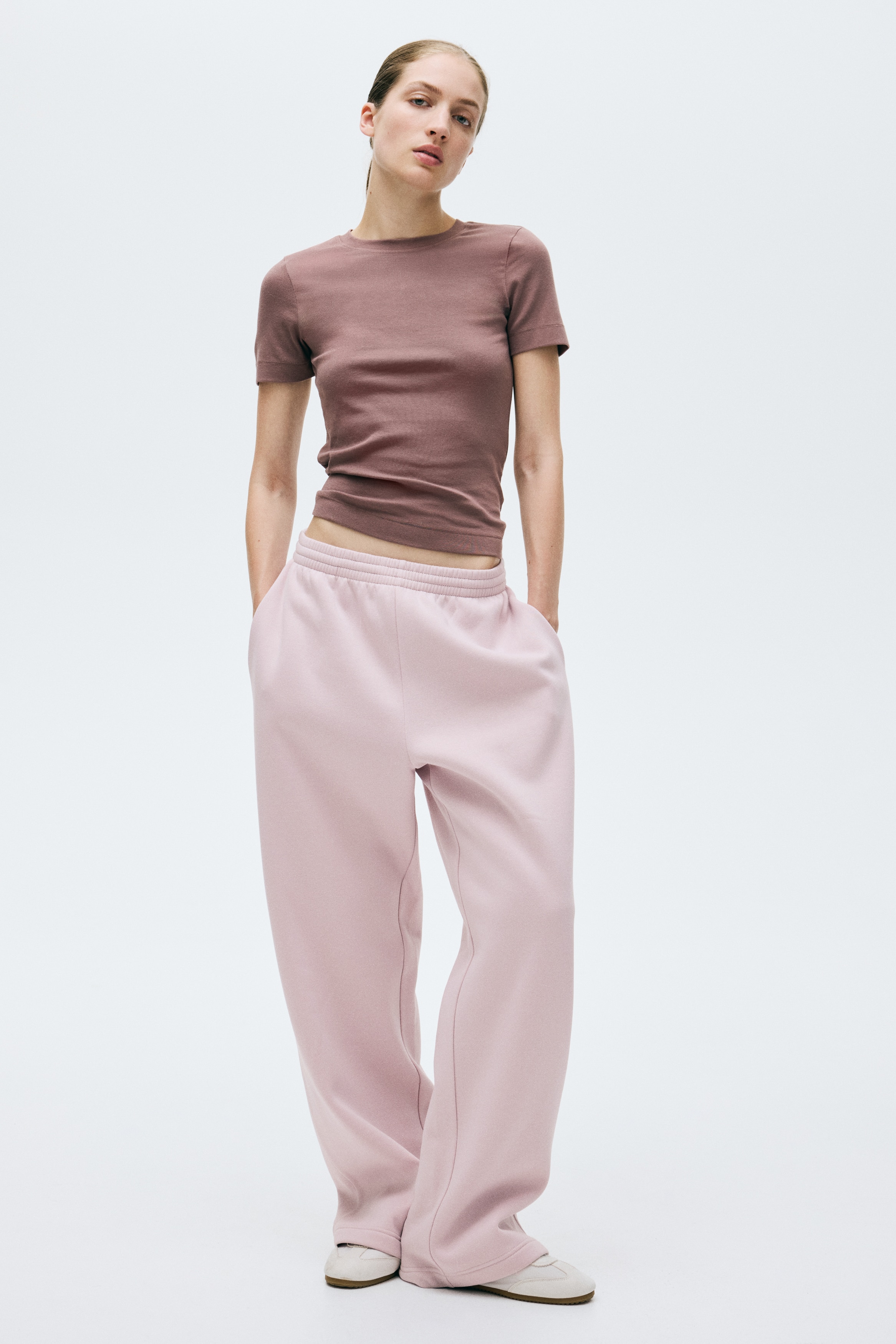 Light pink sweatpants are displaying an elasticated smocking at the waist and visible side pockets. They are featuring straight legs and are presented from a front angle.