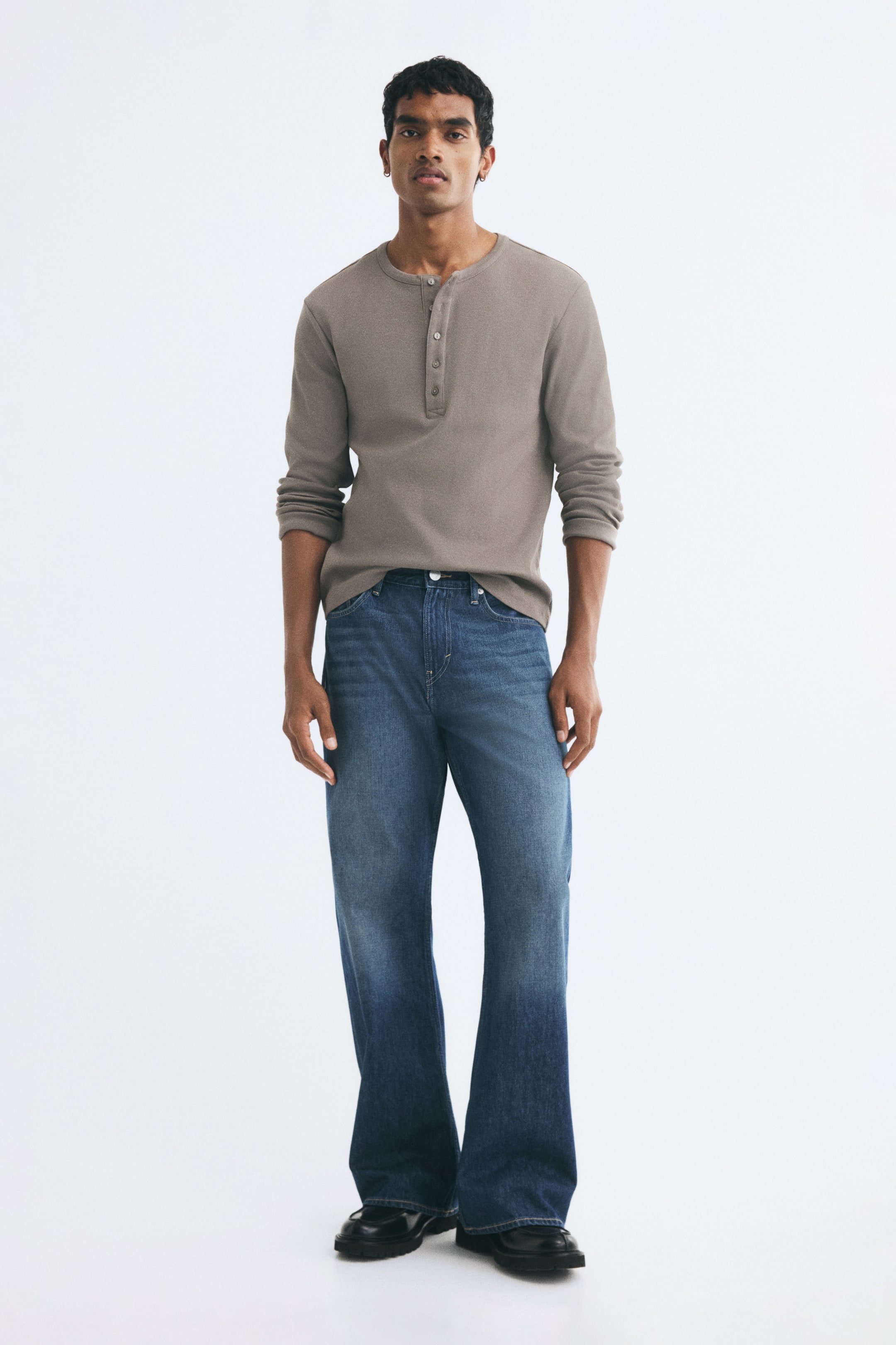 View larger image: Blue denim jeans featuring a relaxed fit, regular waist, and gently flared legs are worn by the model. The medium-wash jeans are styled with a long-sleeve taupe henley shirt and black shoes.