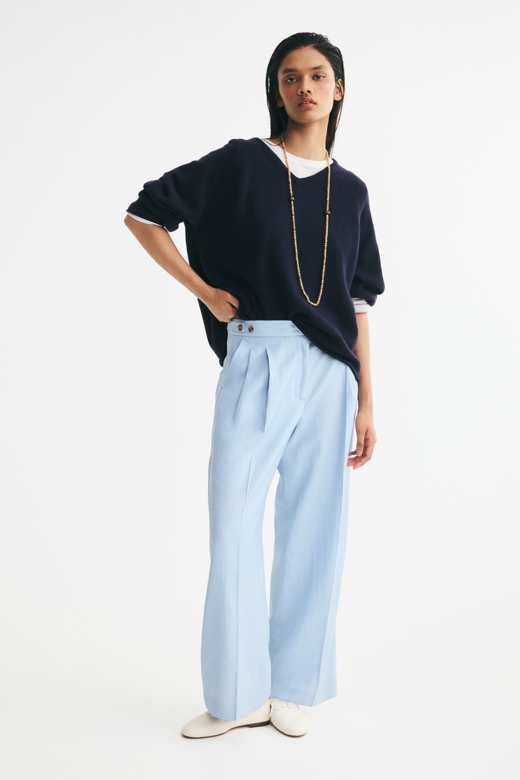View larger image: Light blue pleated trousers, featuring a wide-leg silhouette and a high waist with side button details, are being worn by the model. They are styled with a dark blue V-neck sweater layered over a white T-shirt, a long gold beaded necklace, and white flat shoes.