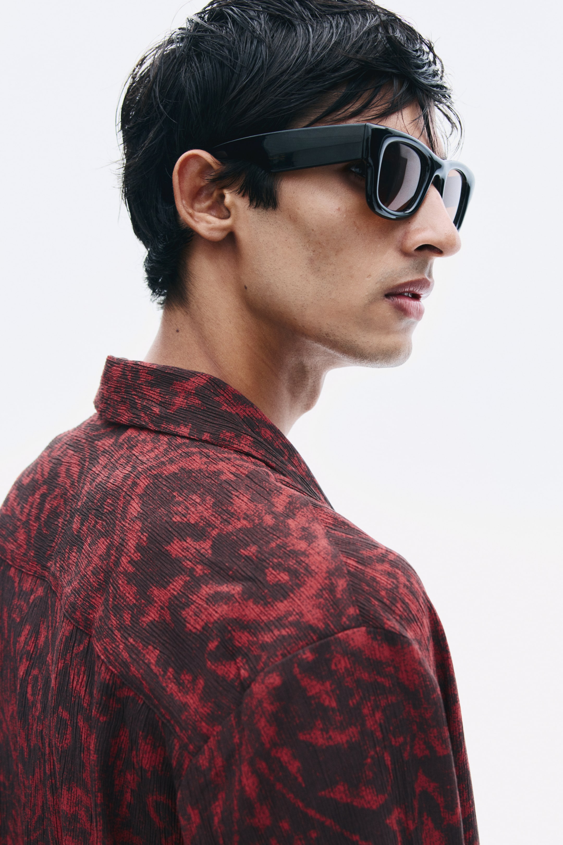 View larger image: A red and black abstract-patterned shirt in a textured weave is being worn by the model. The shirt features a resort collar and is styled with black sunglasses.
