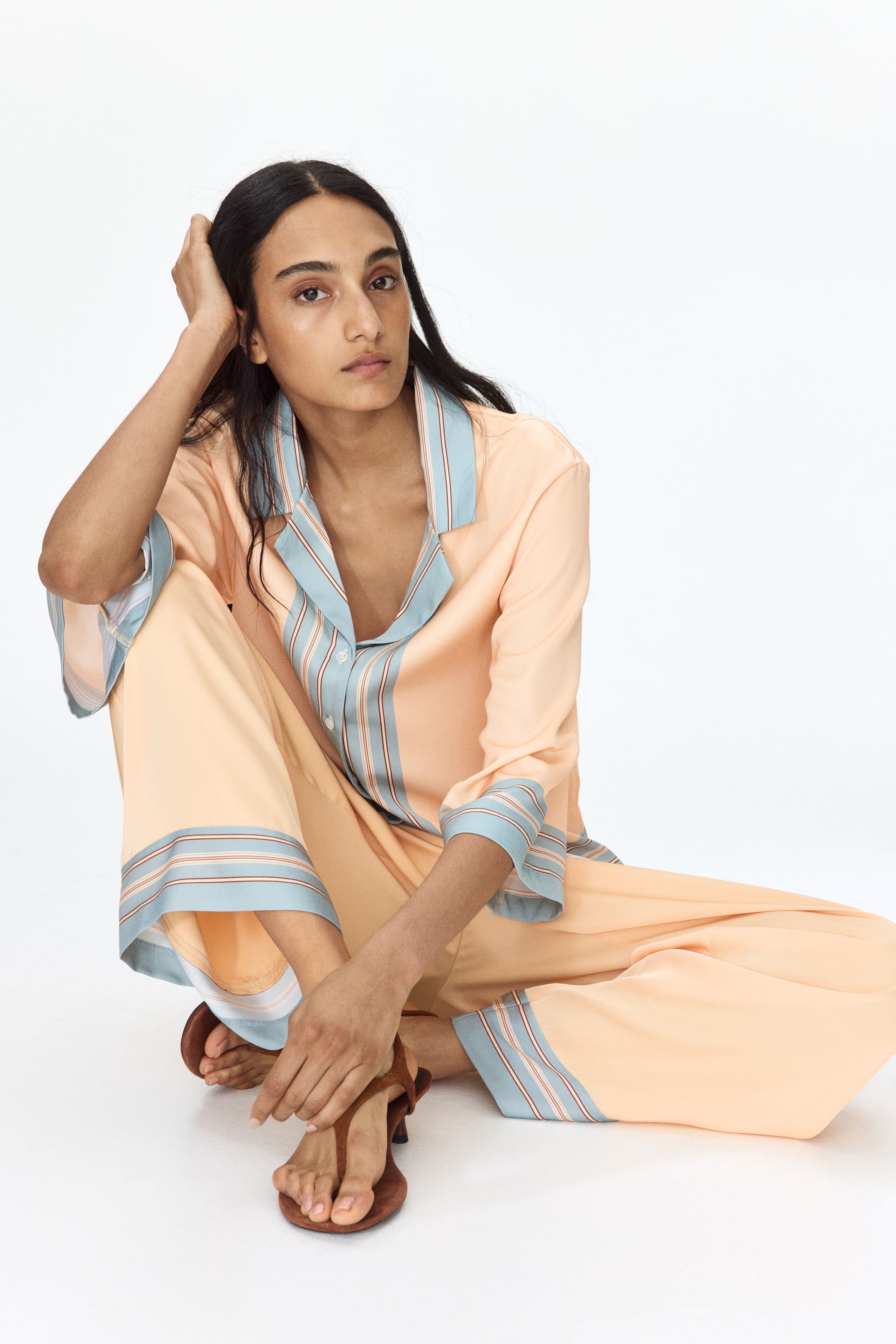 View larger image: The peach pajama set is donned, showcasing a long-sleeved top with notch lapels and wide-leg trousers. The top's collar, cuffs, and the trouser hems are adorned with a light blue trim, highlighted by slender brown, white, and light blue stripes, reflecting the set's laid-back fit and design.