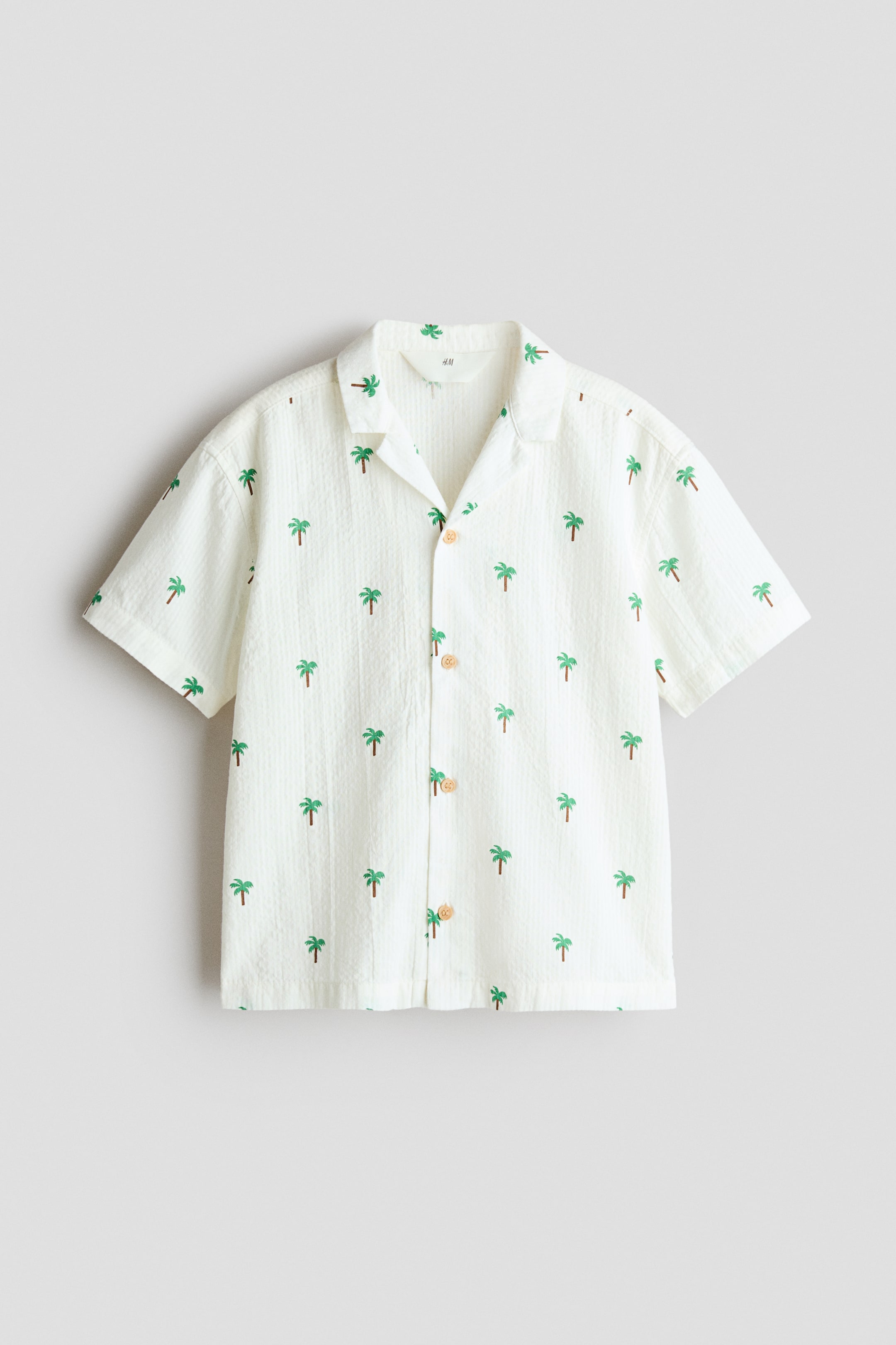View larger image: A soft cotton seersucker off-white loose-fit shirt is displayed facing front. It showcases a pattern of small green palm trees, a resort collar, light brown buttons down the front, dropped shoulders, and short sleeves.