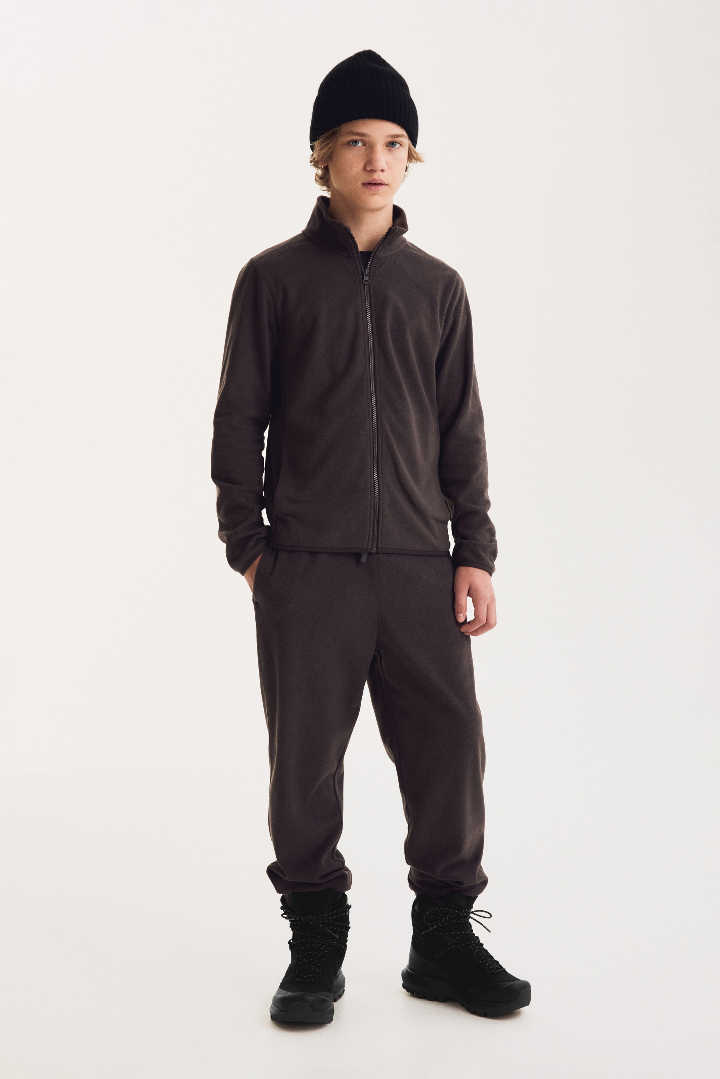 Un pantalon de survÃªtement en polaire marron foncÃ© est suspendu, face avant, avec une taille Ã©lastiquÃ©e et un cordon de serrage assorti rÃ©glable. Le pantalon prÃ©sente une coupe dÃ©contractÃ©e avec des poches latÃ©rales et des chevilles resserrÃ©es par des poignets Ã©lastiquÃ©s.
